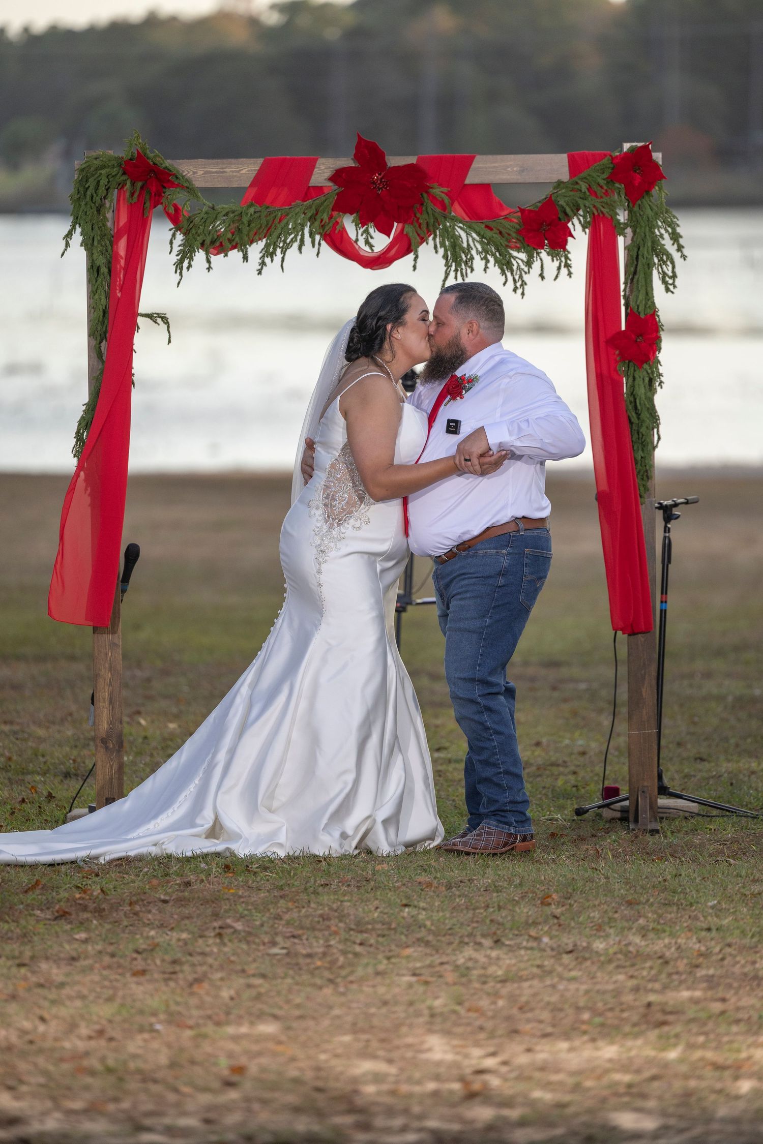 A romantic outdoor wedding ceremony under a rustic wooden arch decorated with red fabric and greenery.
