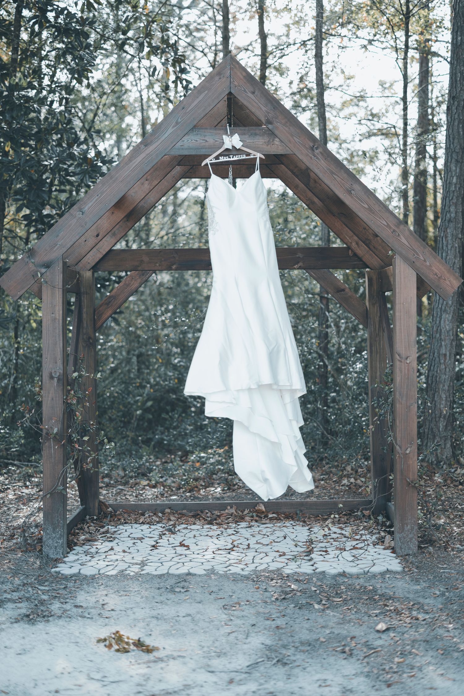 A white wedding dress hangs from a wooden arch in a forest setting creating an ethereal outdoor scene.