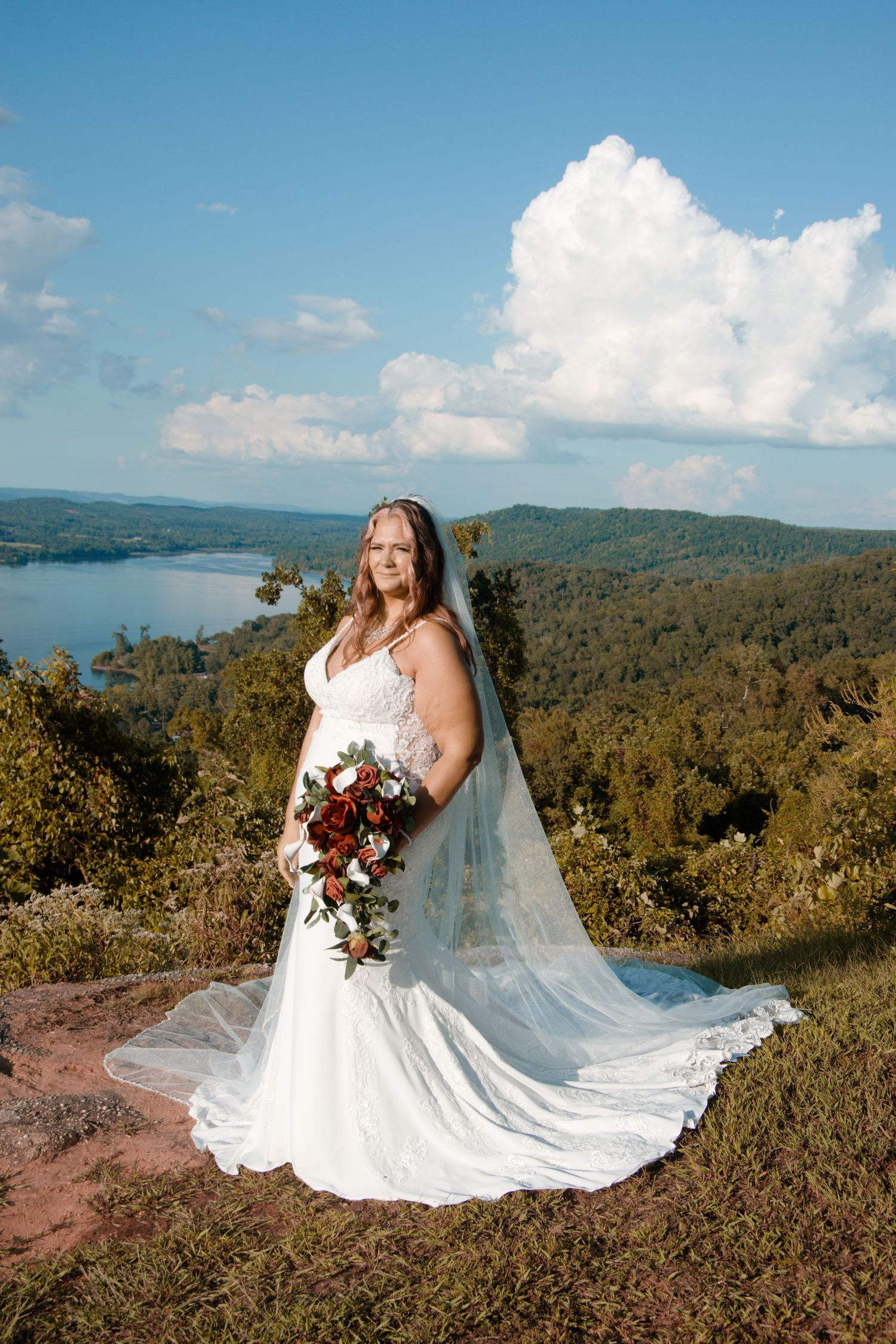 A scenic mountaintop wedding photo shows a flowing white dress and veil against a backdrop of rolling hills and blue sky.