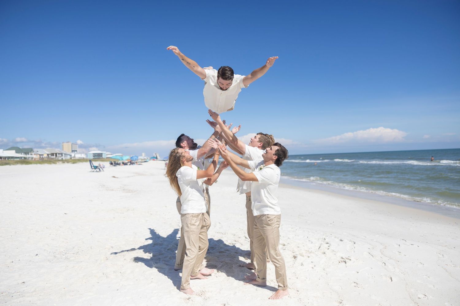 Wedding party celebrates on a sunny beach with arms raised while ocean waves crash in the background.