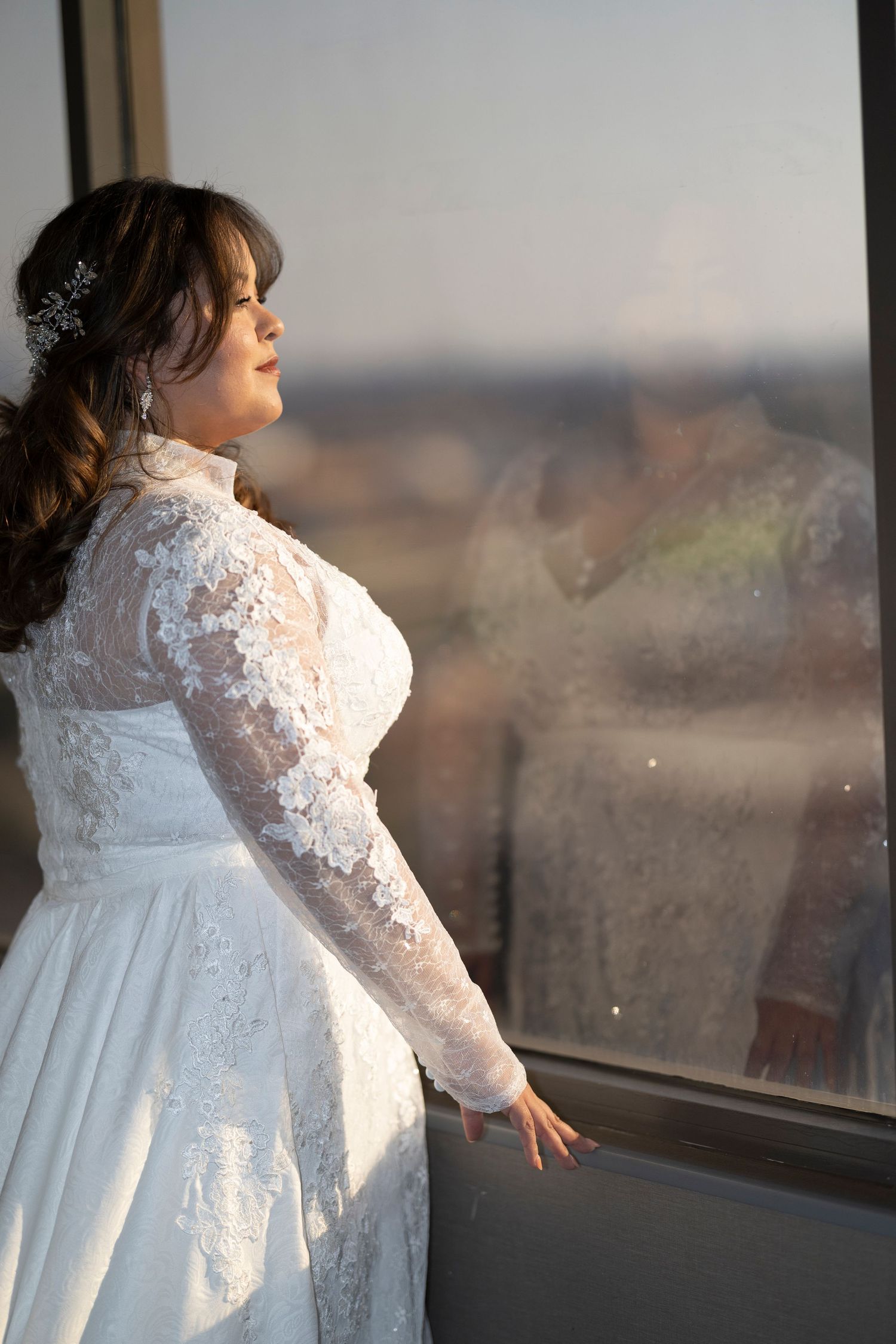 A person in a white lace wedding dress poses near a window during golden hour, creating a dreamy silhouette effect.