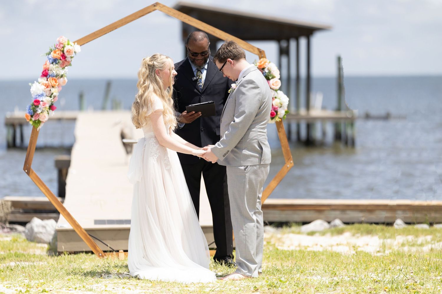 A couple exchanges vows during a waterfront wedding ceremony under a wooden arch decorated with flowers.