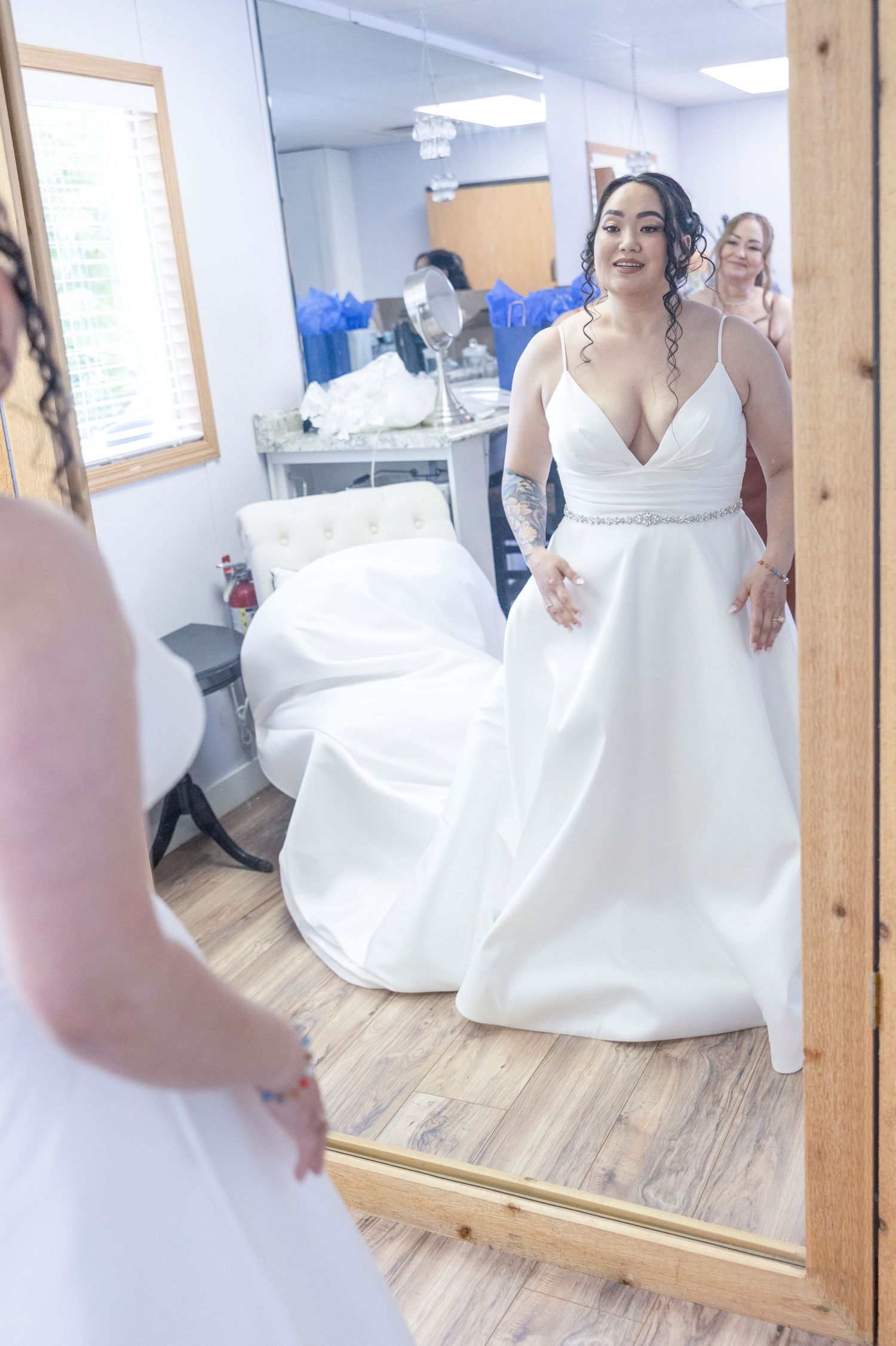 A bride in a white wedding gown looks at her reflection in a bridal boutique fitting room mirror.