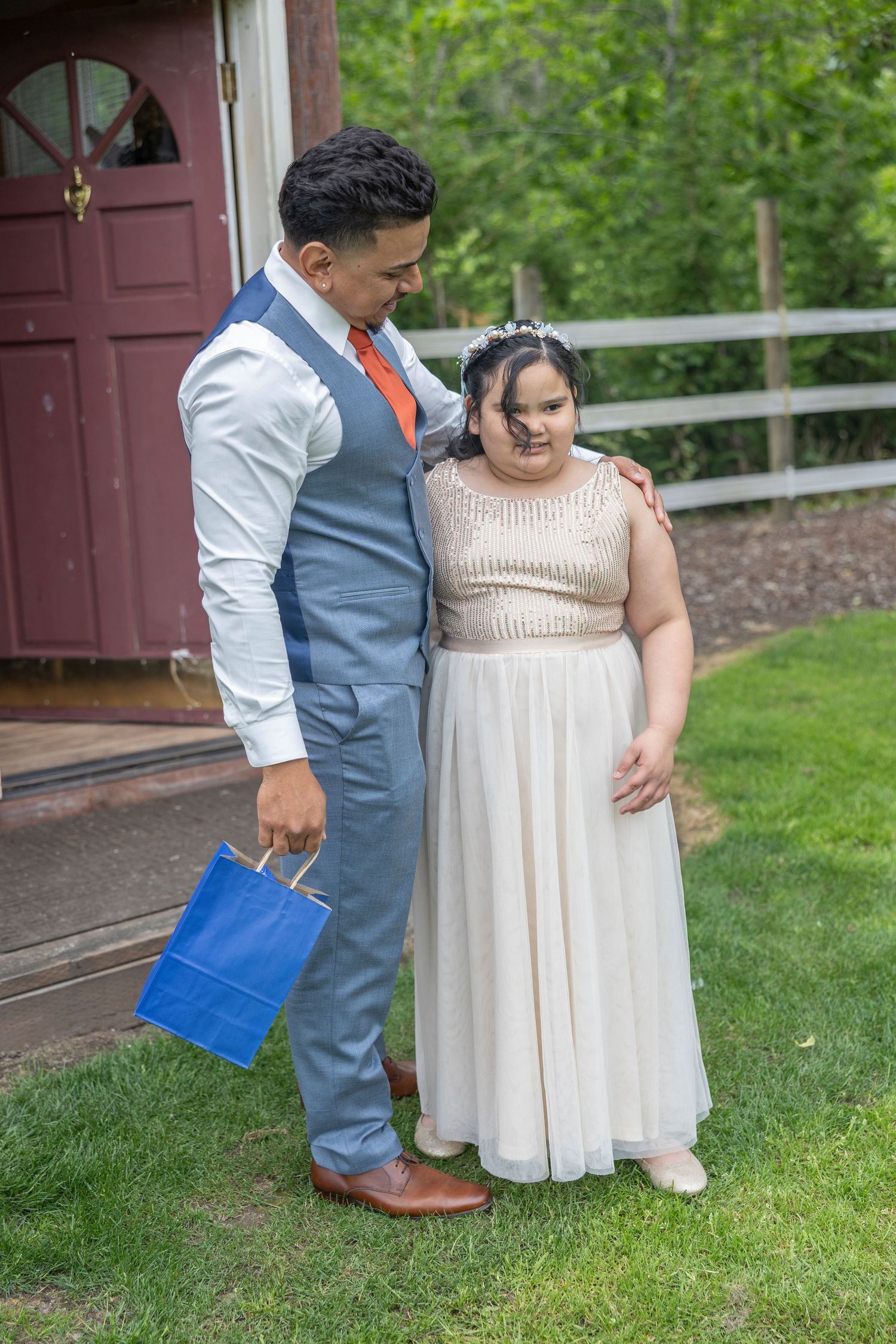 A couple in formal attire stands together on grass outside a building, sharing an intimate moment.
