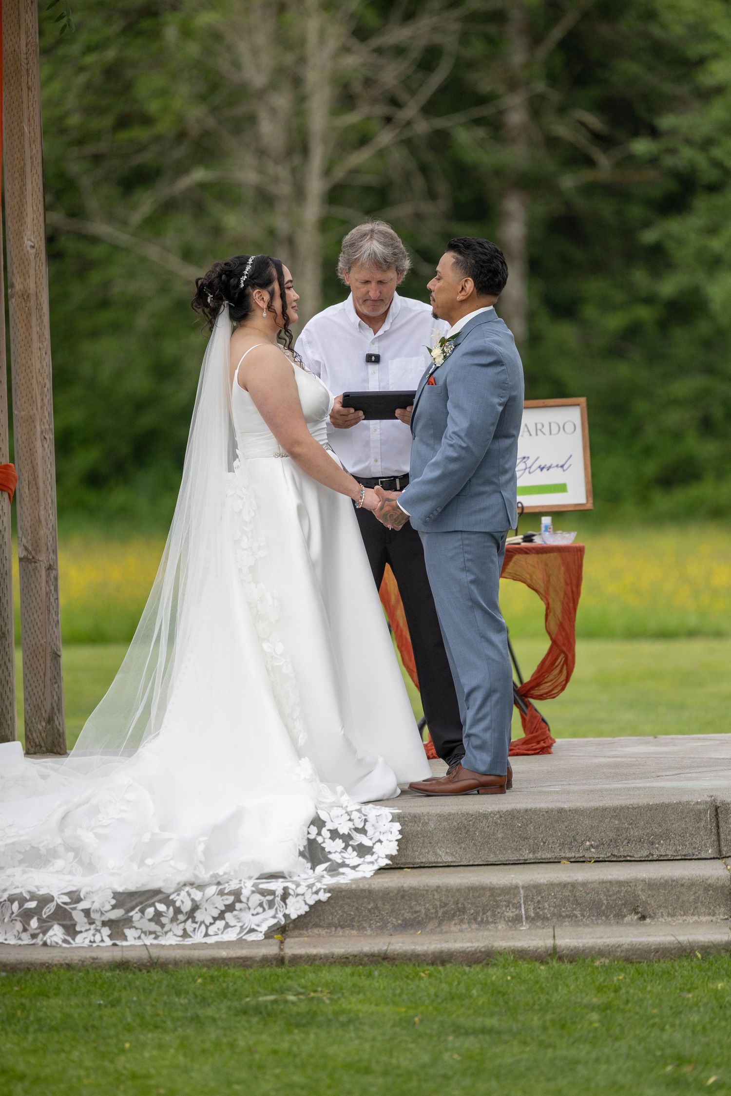Wedding ceremony taking place under a rustic wooden pavilion surrounded by green grass and trees.