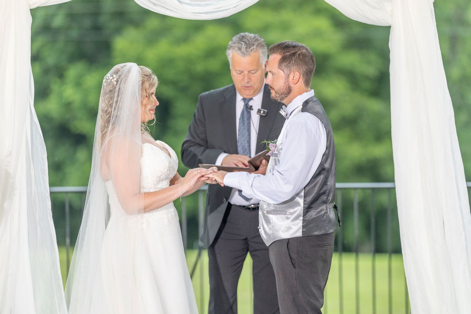 Wedding ceremony takes place outdoors on a scenic deck with white curtains and green trees in the background.
