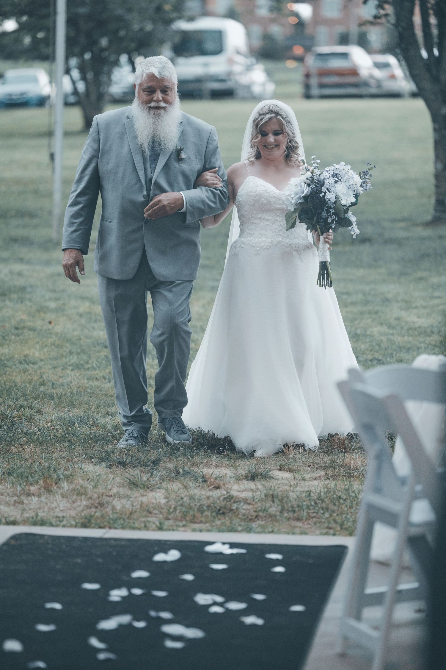 A wedding processional shows a bride in a white dress being escorted down an aisle with scattered flower petals.