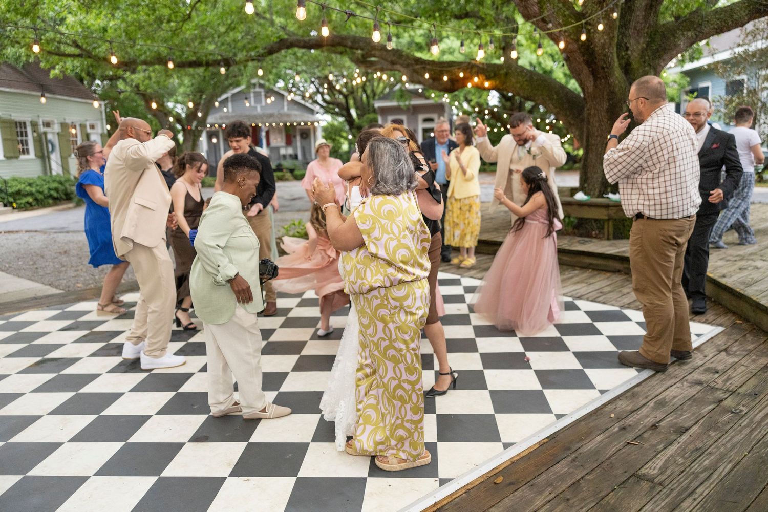 People dancing on a black and white checkered outdoor patio under string lights and tree canopy during summer event.