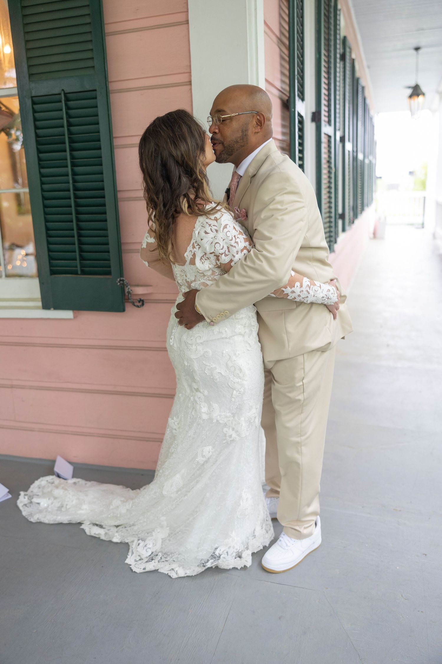 A couple shares a romantic kiss on a porch with green shutters and pink walls wearing elegant white wedding attire.