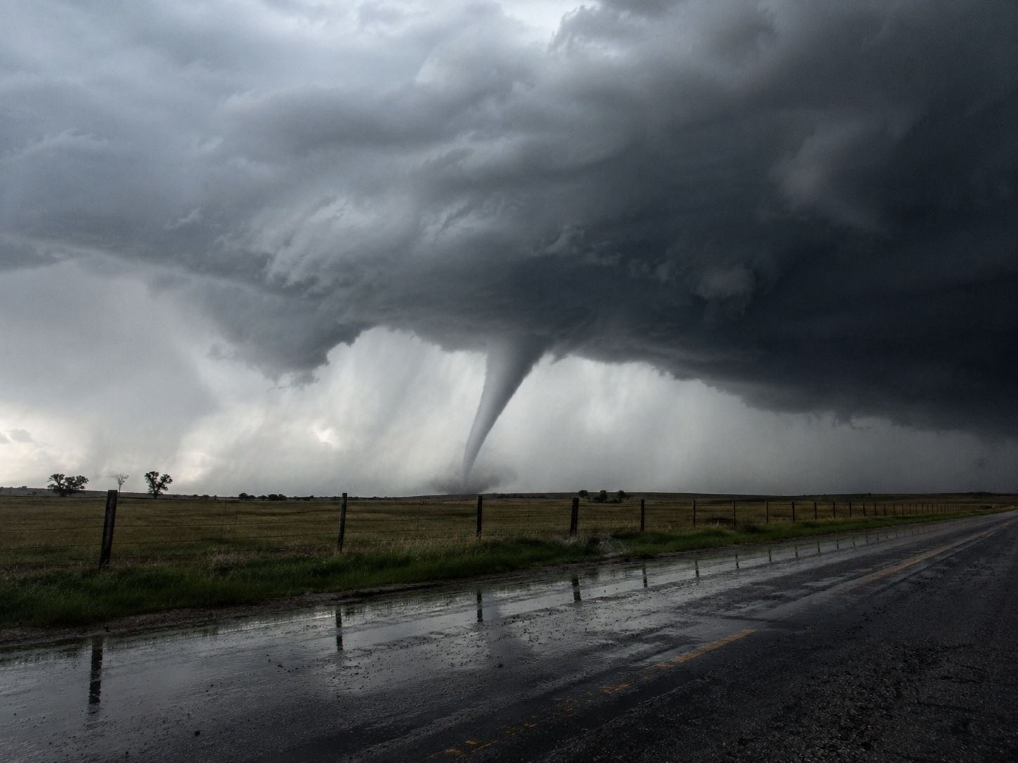 A massive tornado touches down on a rural road under dark storm clouds in an open field.
