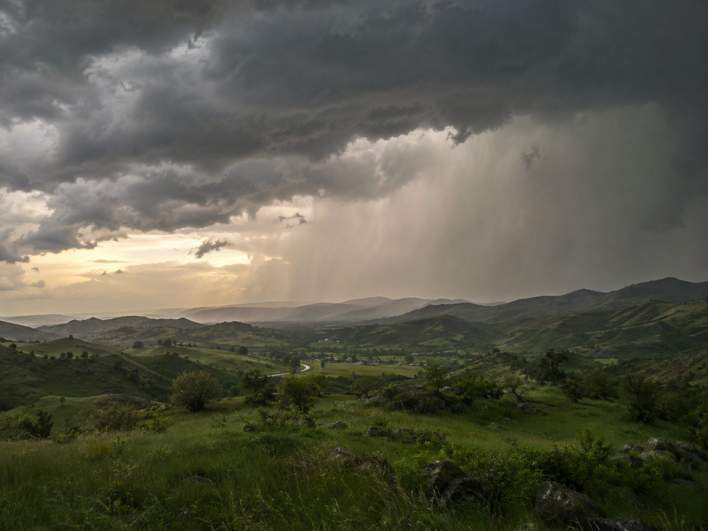 Dramatic storm clouds over lush green rolling hills with rain curtains and golden sunset light breaking through the sky.