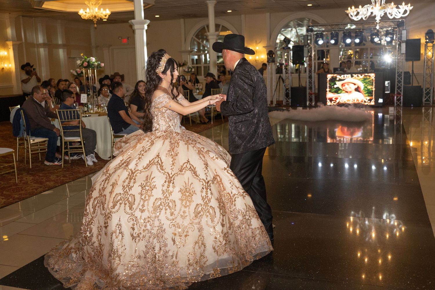 A young woman in a gold quinceañera gown dances with a partner in a elegant ballroom celebration.