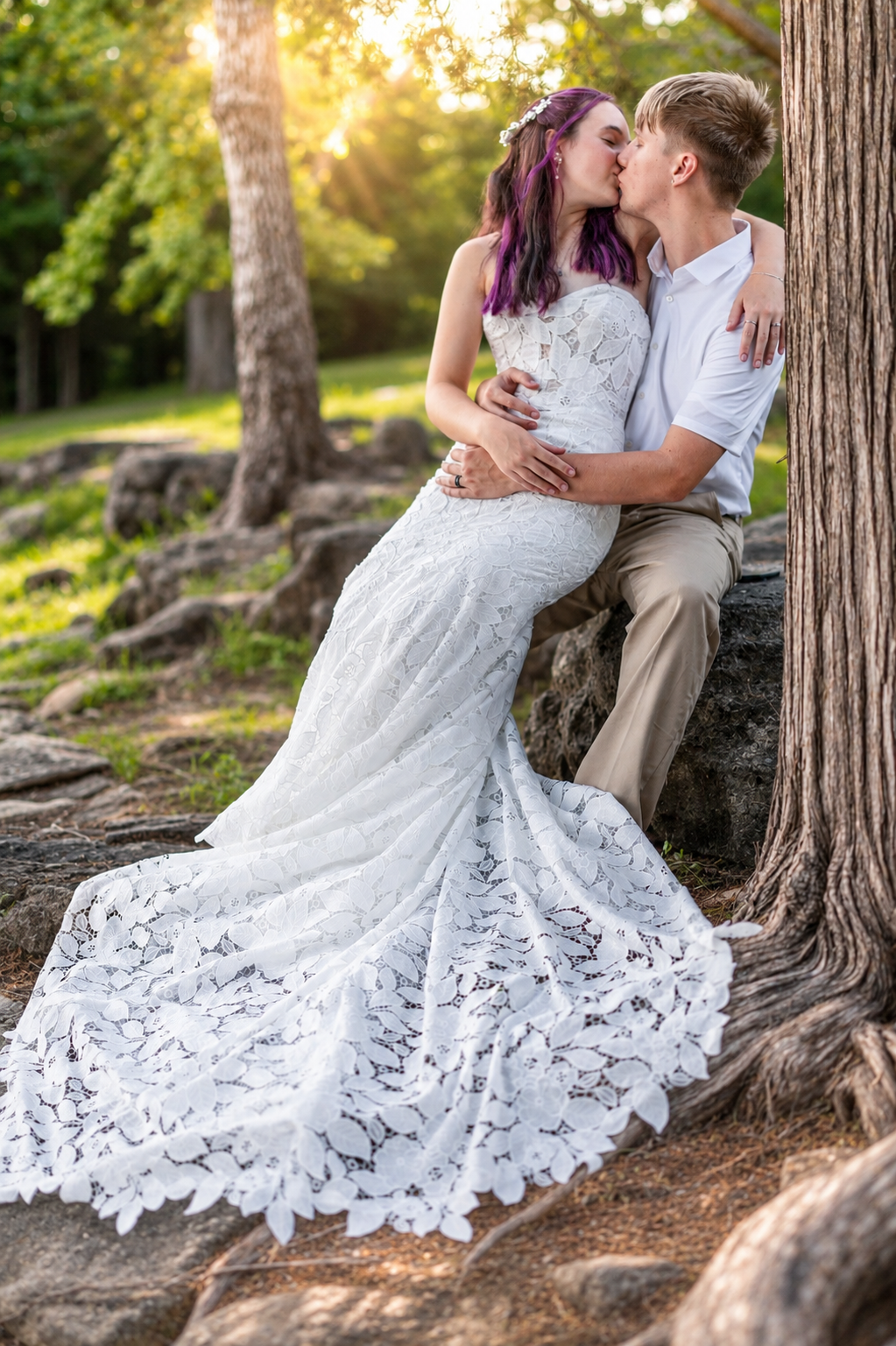 Bride in white lace mermaid gown kissing groom outdoors on rocks surrounded by trees in golden sunset light.