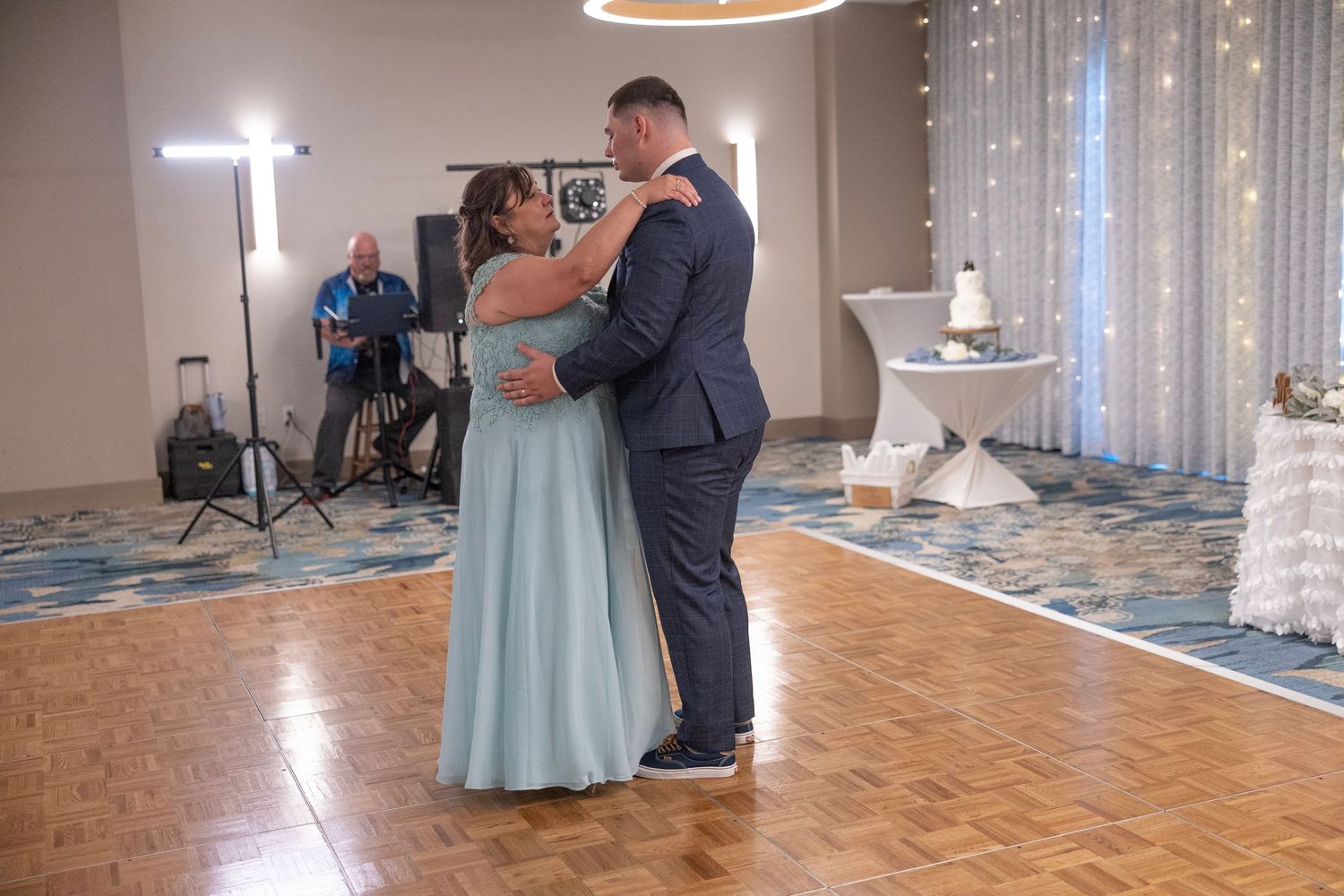 Couple shares first dance at wedding reception on wooden dance floor with fairy lights and musician in background.