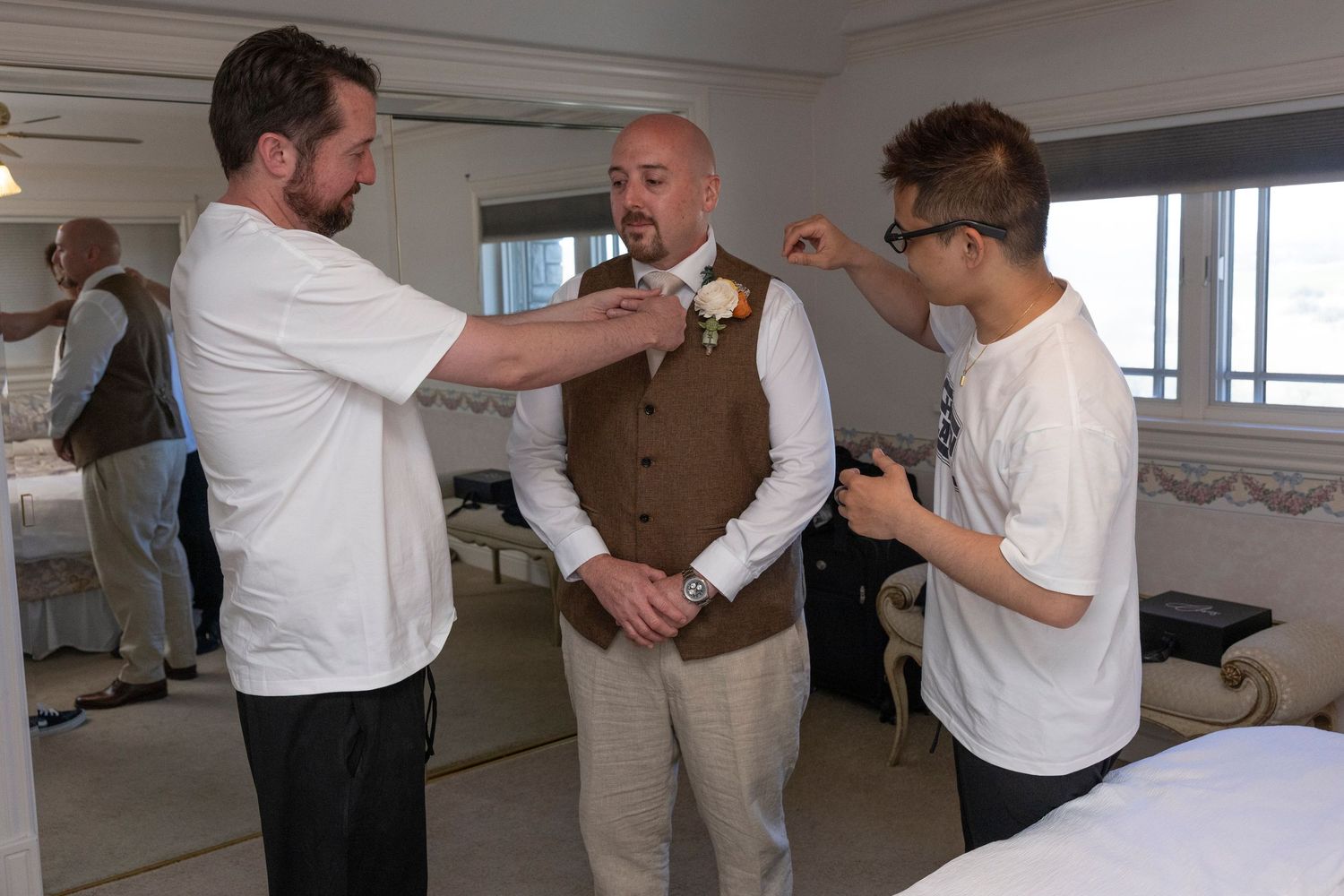 Groom being helped with boutonniere by two men in a hotel room before wedding ceremony.