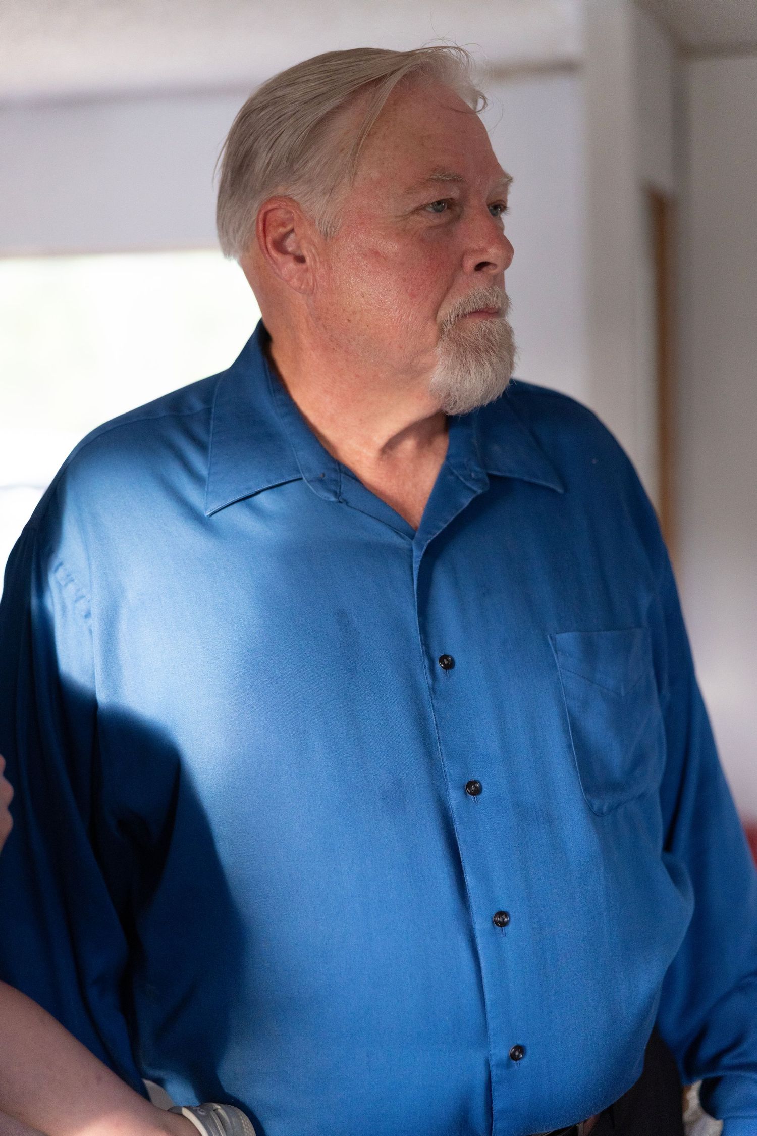 Older man with white hair and beard wearing a blue button-up shirt, standing indoors with a thoughtful expression.