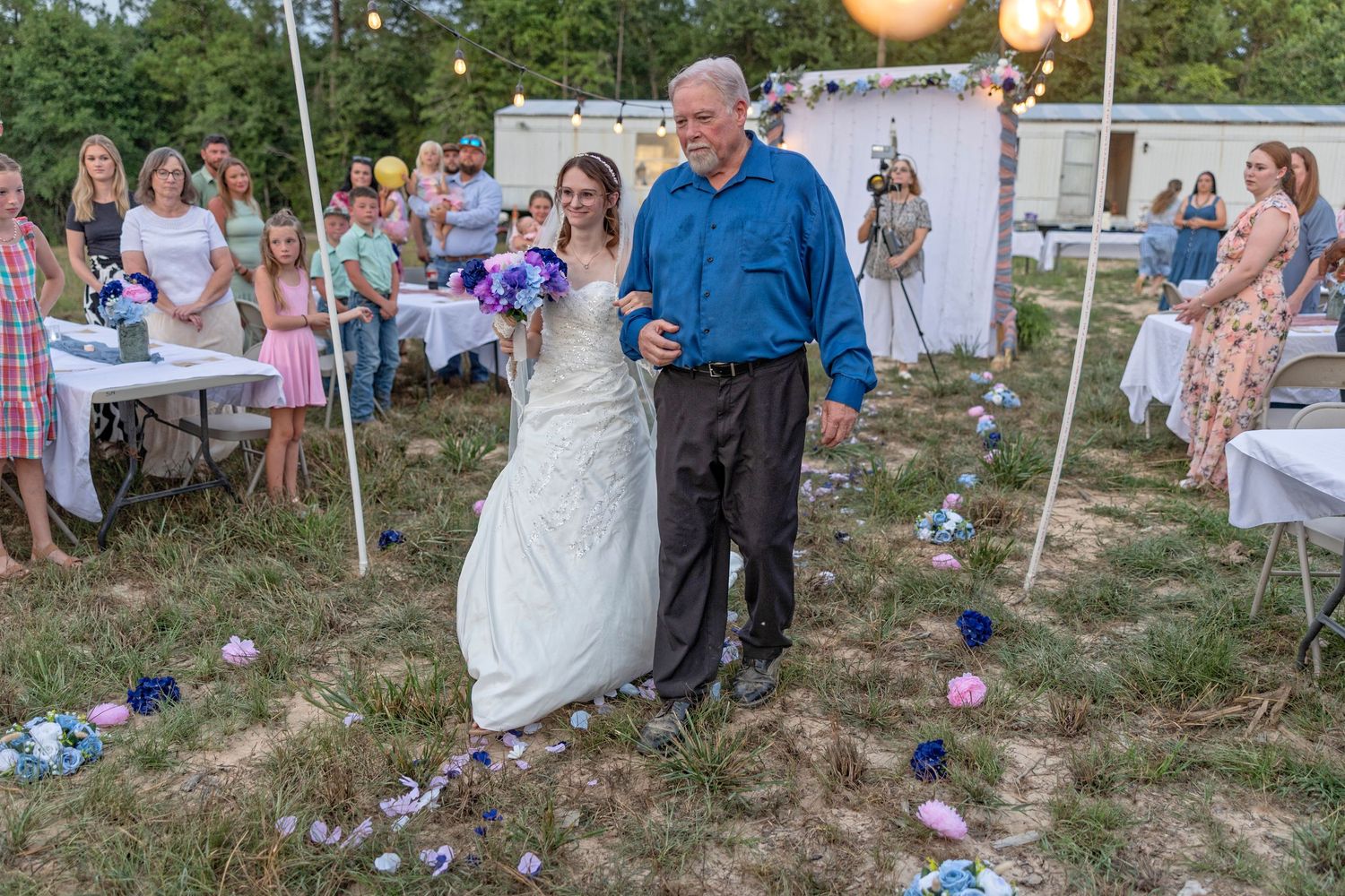 Bride in white gown walks down outdoor aisle with older man in blue shirt at rustic backyard wedding.