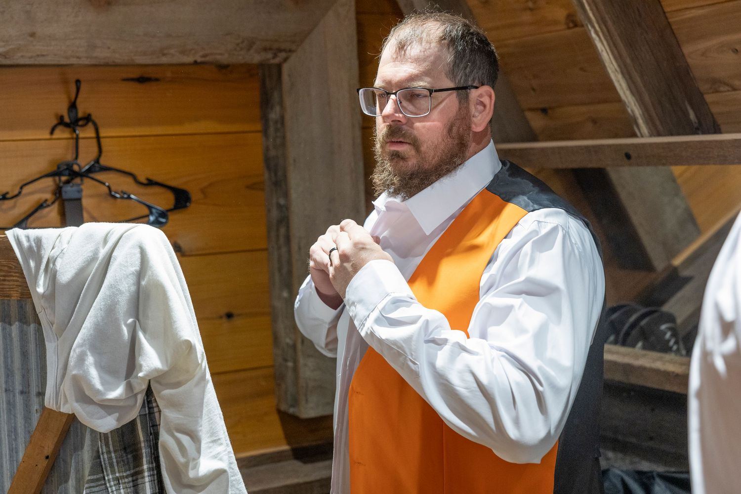 Man in orange vest and white shirt adjusting his tie in a rustic wooden setting.