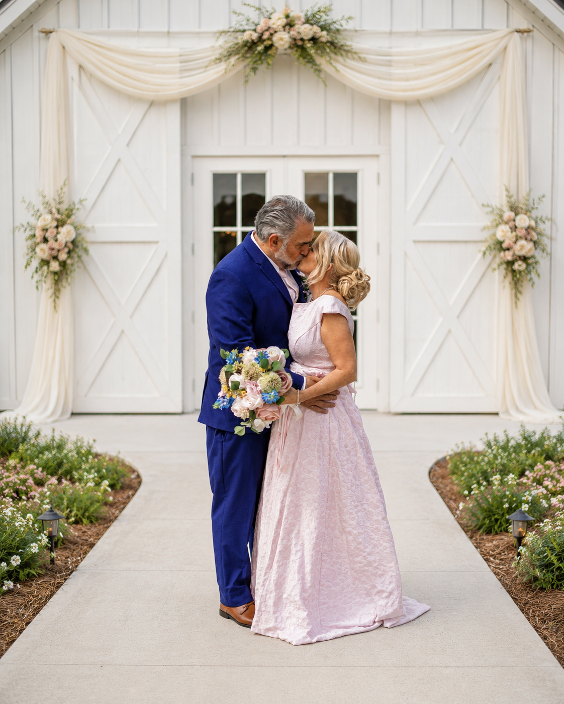 Couple sharing a romantic kiss in front of a white barn decorated with floral arrangements and draped fabric.