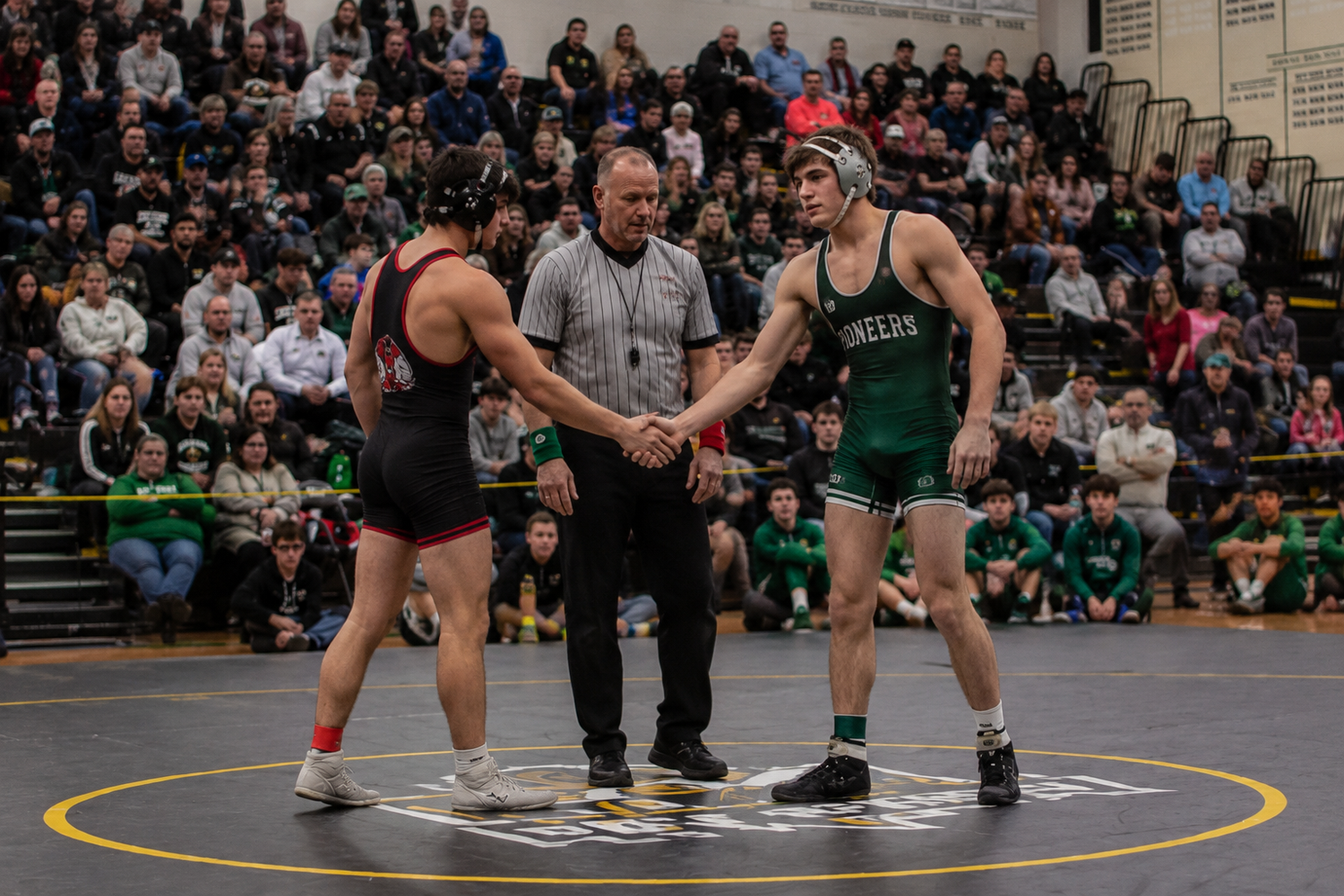 A referee oversees a handshake between two wrestlers on a yellow-bordered mat at a wrestling tournament.