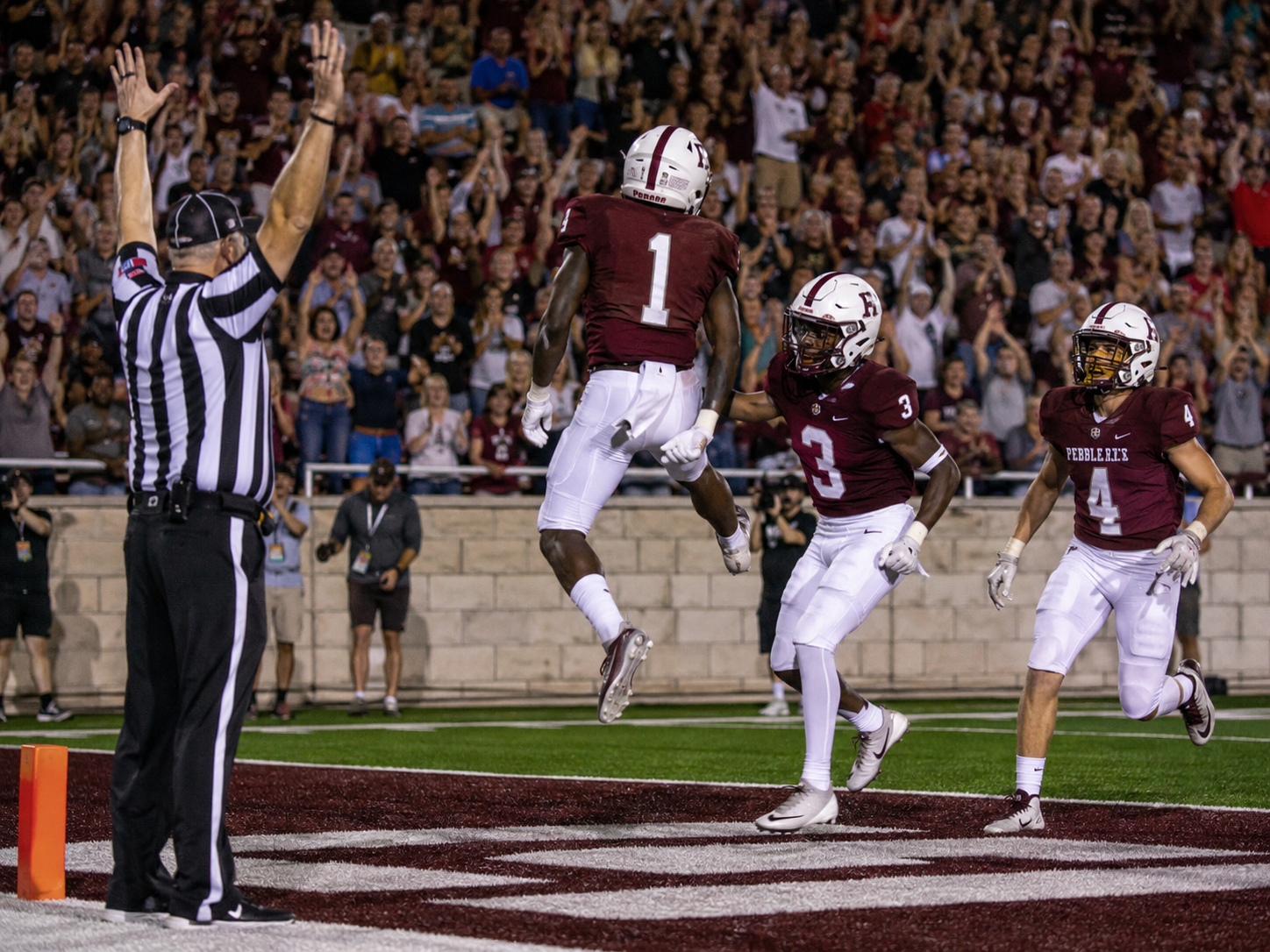 A college football player leaps in celebration after scoring a touchdown as a referee signals the score.