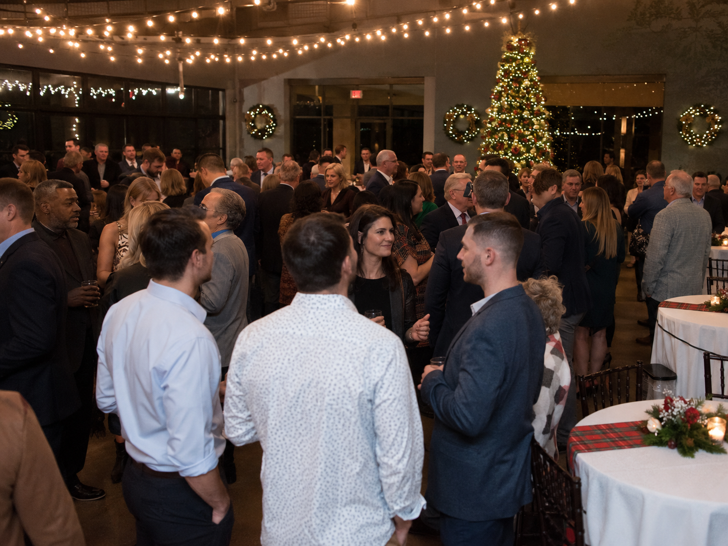 Large holiday party crowd mingles under string lights near a decorated Christmas tree.