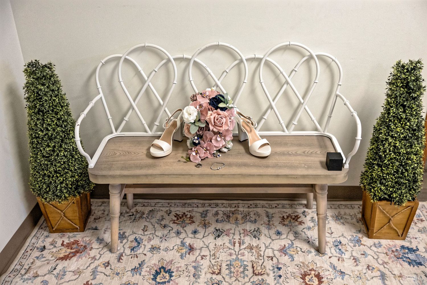 White bench with floral bouquet and bridal heels, flanked by topiary plants on a patterned rug.