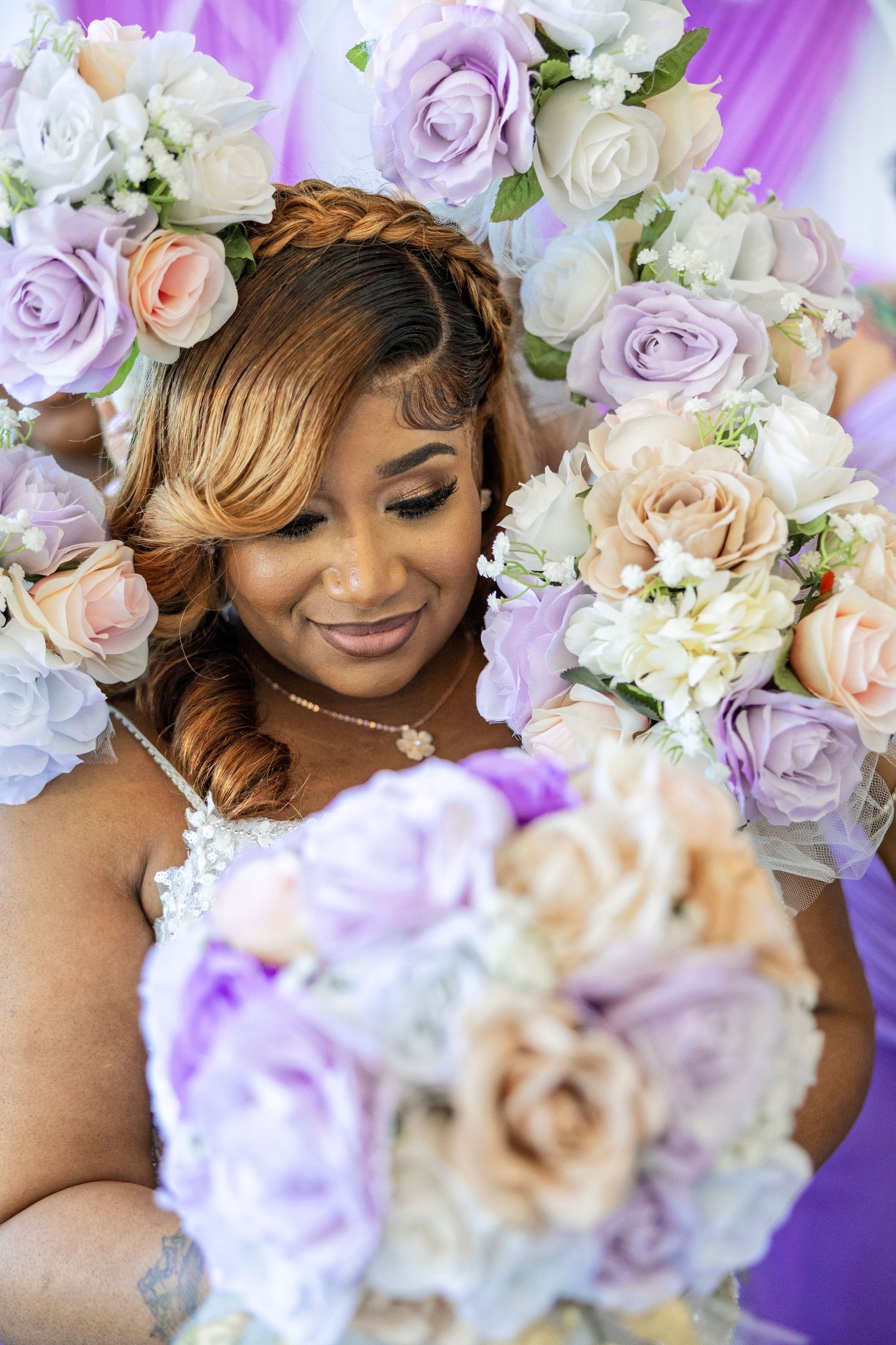 A bride with braided updo smiles softly surrounded by lavender and blush rose bouquets at a wedding ceremony.