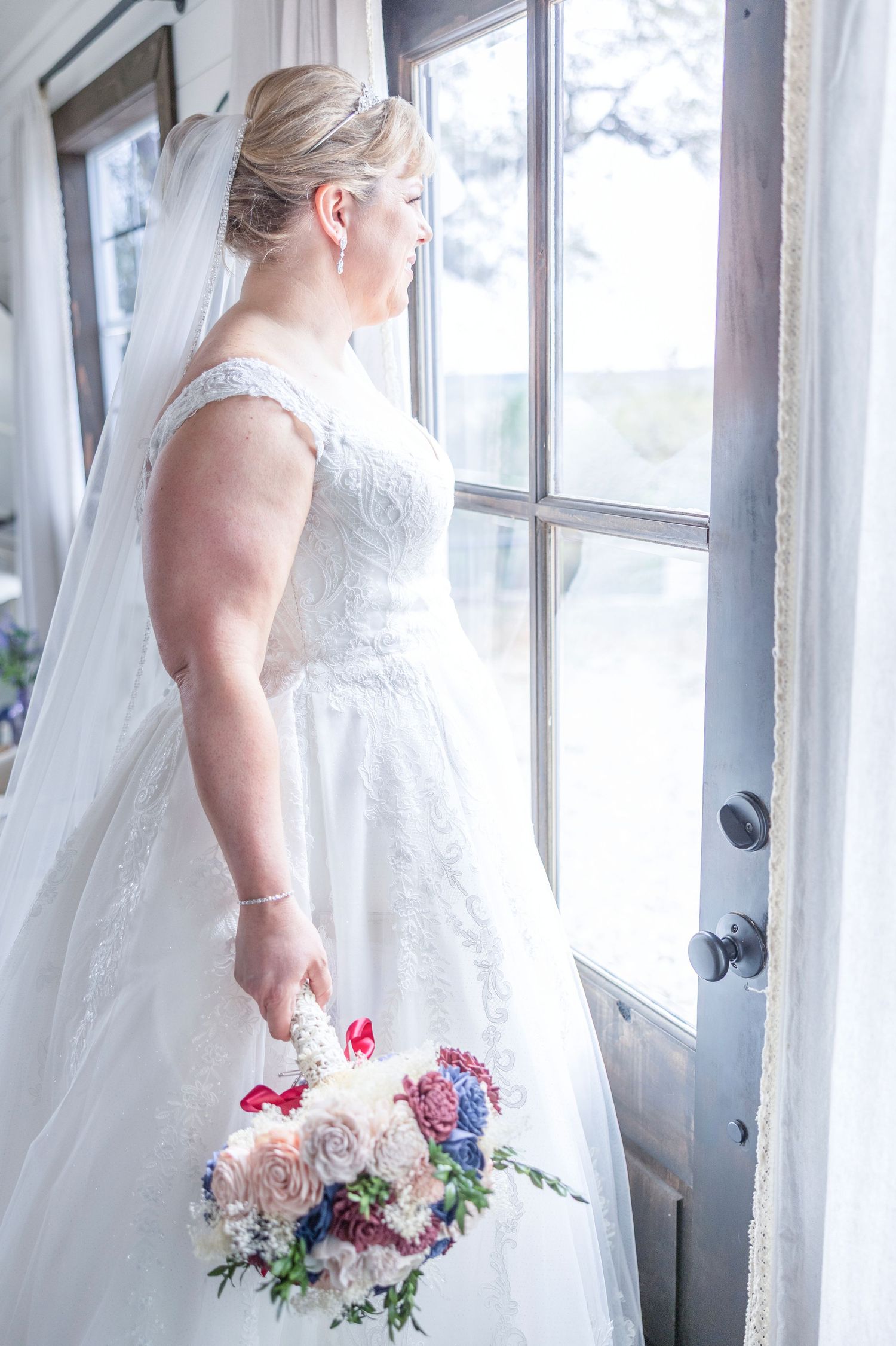 Bride in white lace ball gown holding colorful bouquet, gazing out window with flowing veil.