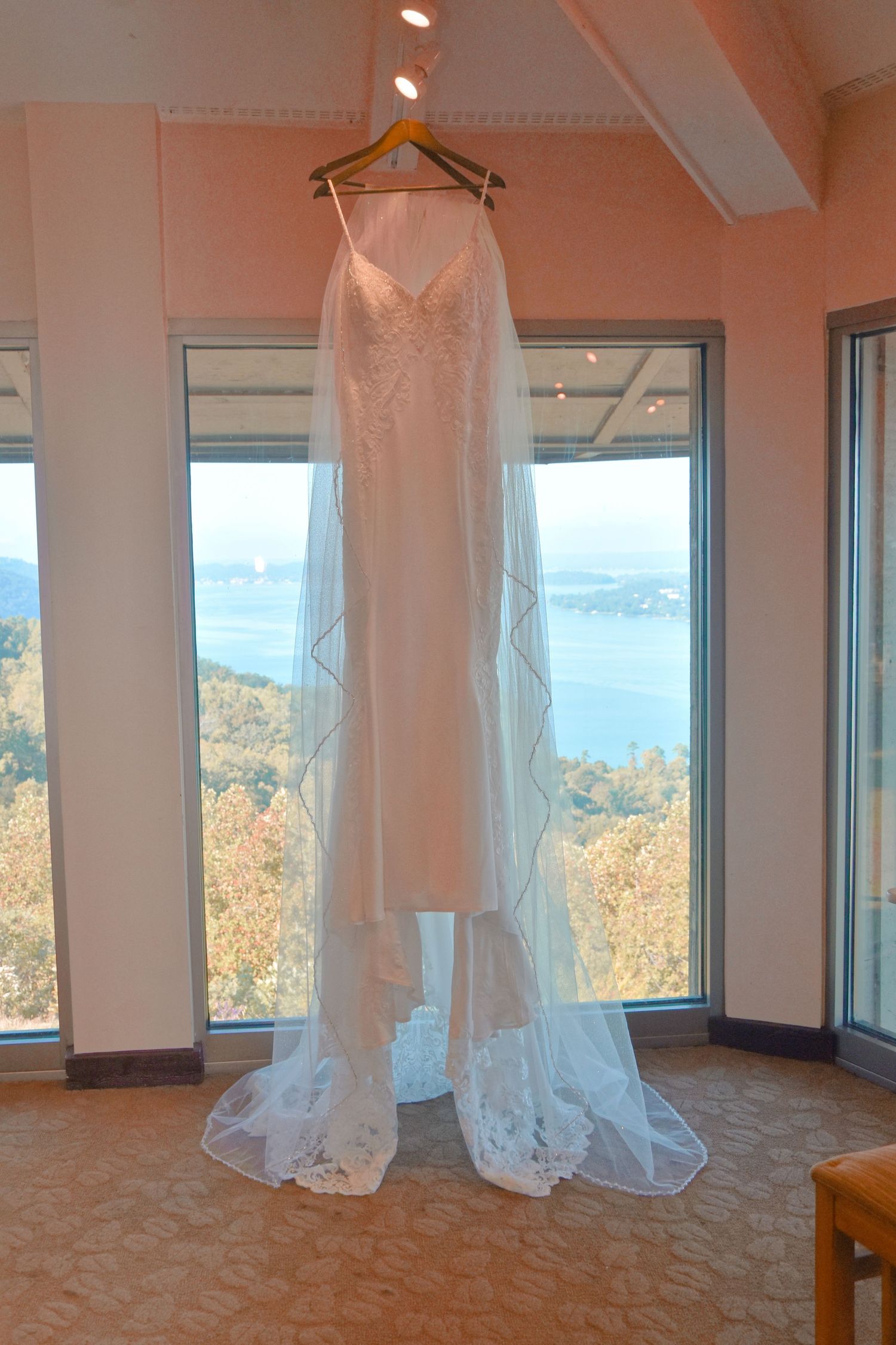 Elegant white wedding dress with lace train hanging in sunlit room with panoramic mountain and lake views.