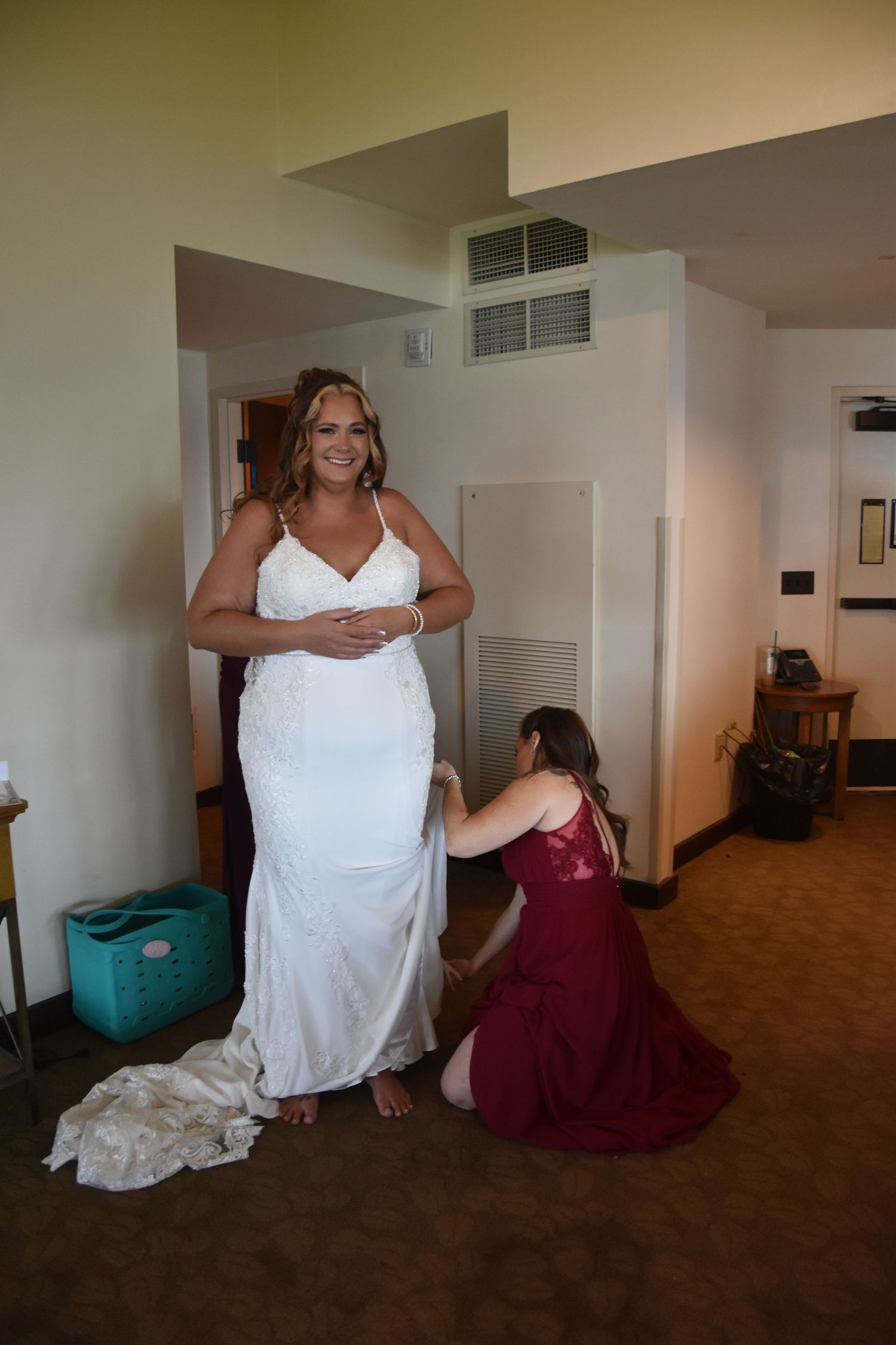 A wedding dress fitting scene shows someone in a white gown while another person adjusts the dress in a carpeted room.