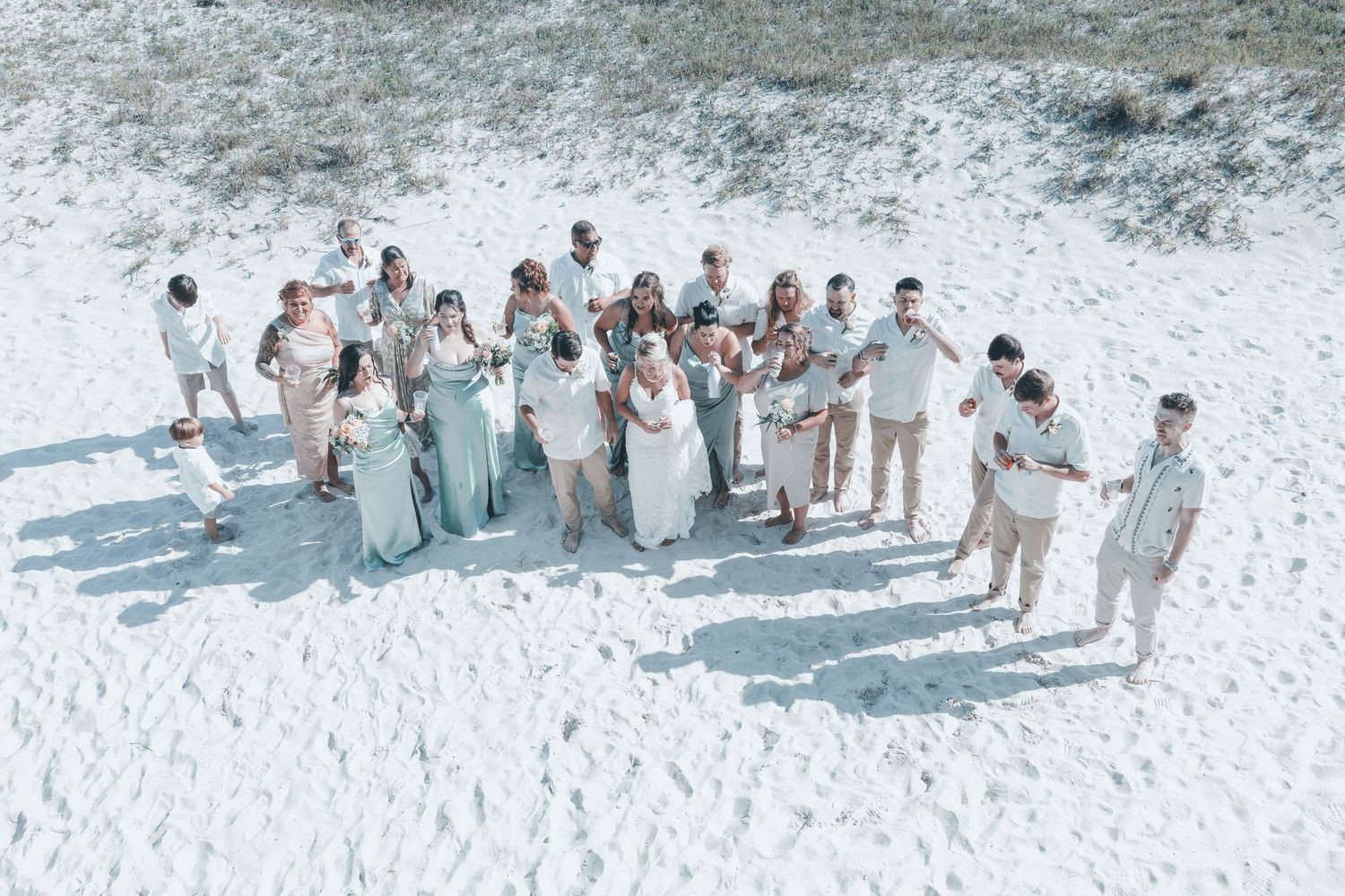 A wedding party in light blue and beige attire poses together on a white sandy beach in a series of group photos.