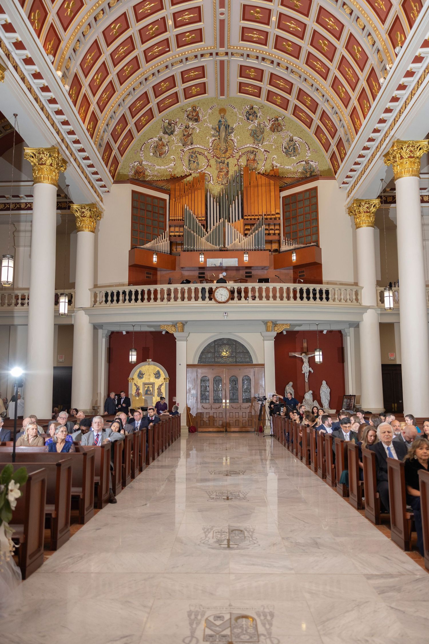Interior view of an ornate church with vaulted ceiling, marble columns, wooden pews and a grand pipe organ at the altar.