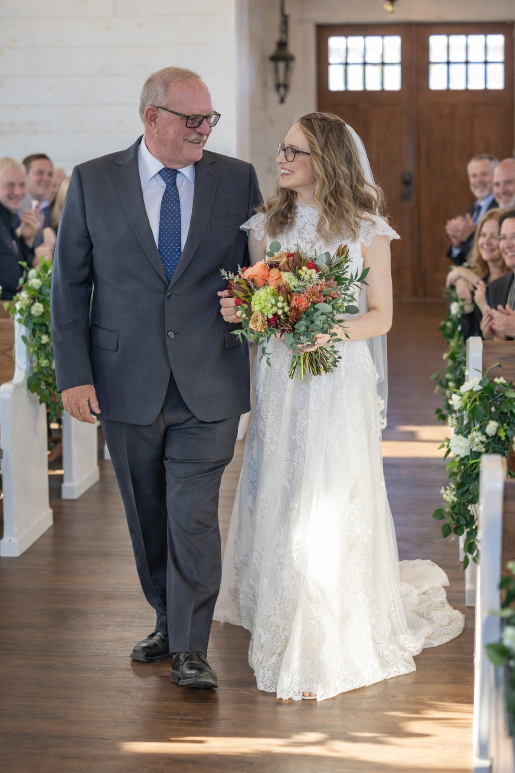 A wedding ceremony processional down a wooden-floored aisle with floral decorations in an elegant indoor venue.