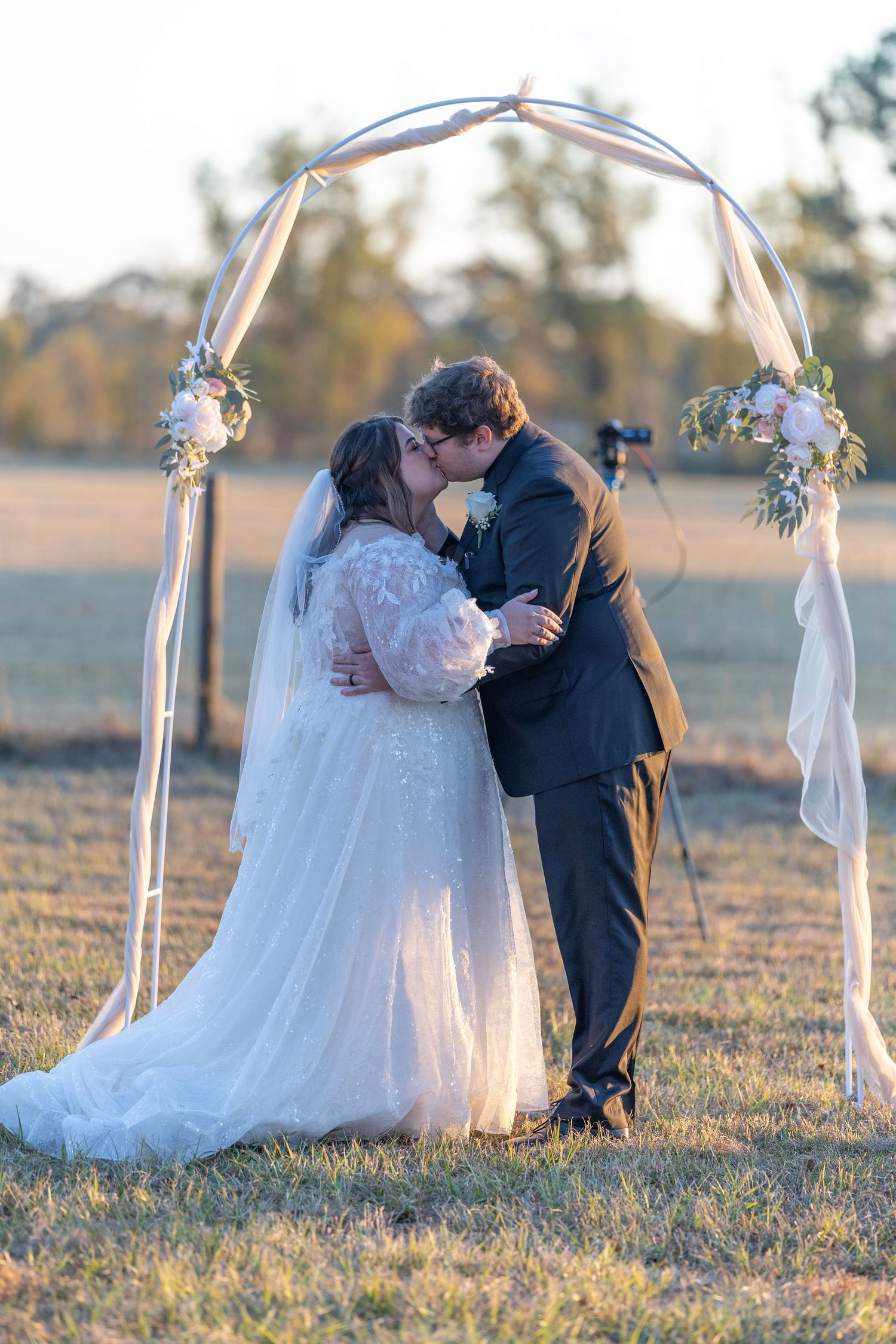 A romantic wedding couple shares intimate moments under a floral arch in a sunlit field at golden hour.