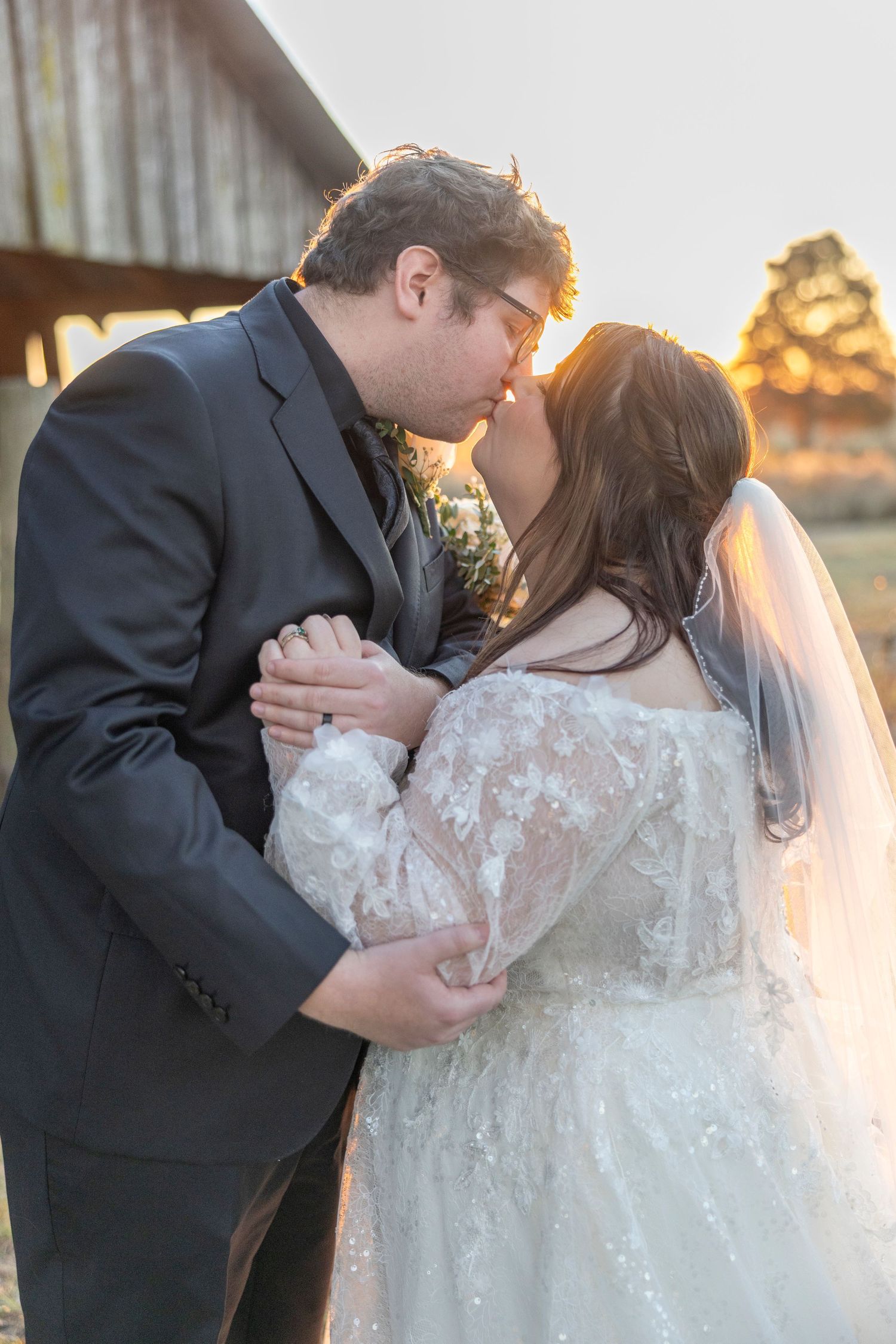 A romantic sunset wedding photo shows a couple sharing a tender kiss in front of a rustic barn.