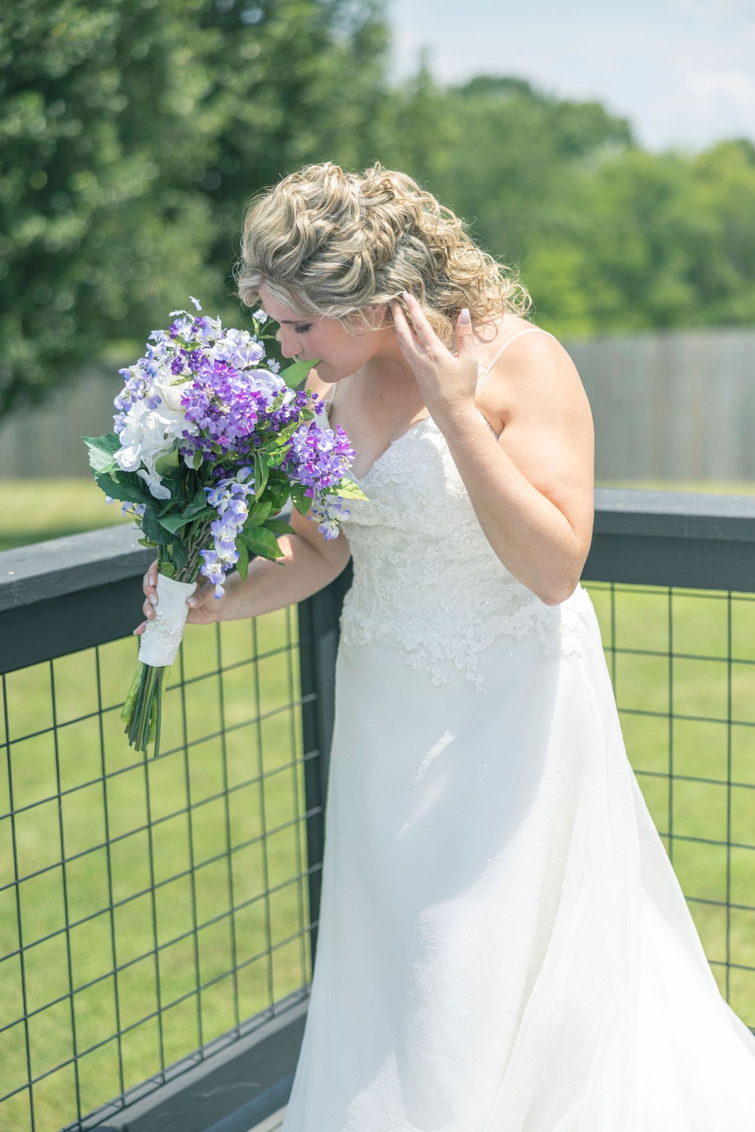 A bride in a white wedding dress holds a purple floral bouquet while standing against an outdoor fence.