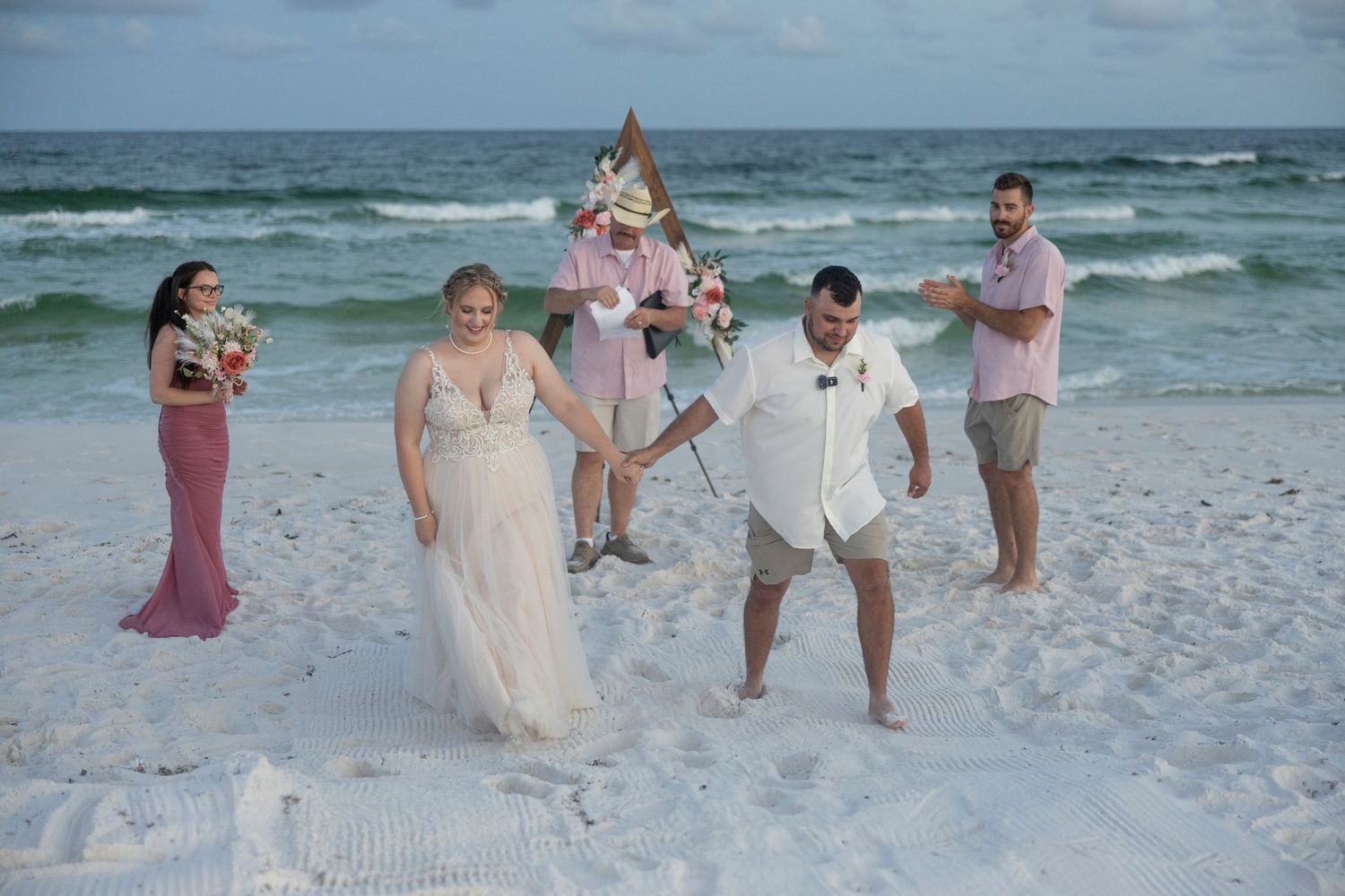A wedding party walks together along a white sandy beach with gentle waves and cloudy skies in the background.