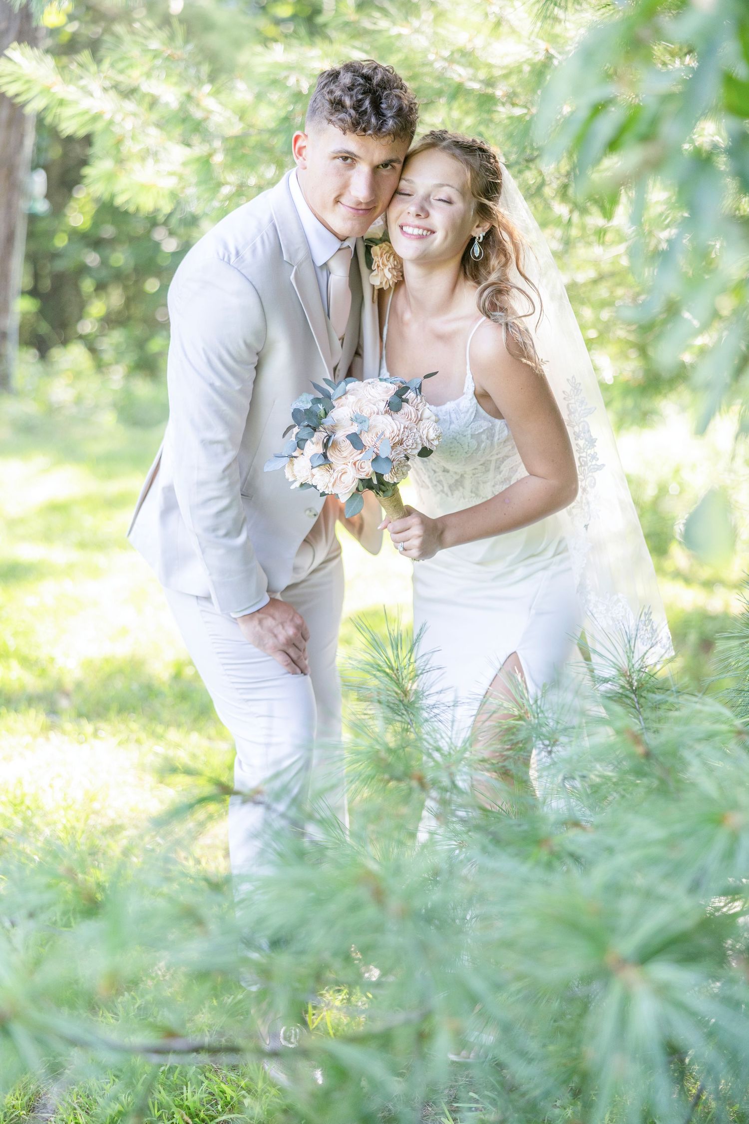 A romantic outdoor wedding photo with a couple in white attire holding a pastel bouquet amid sunlit greenery.