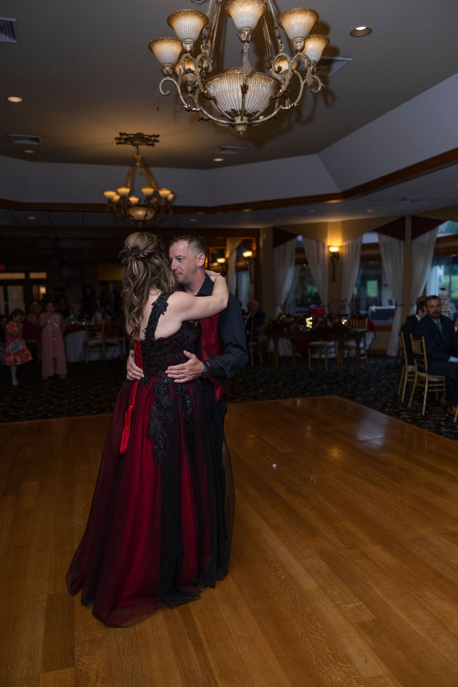 Two people share an intimate dance on a polished hardwood floor under warm chandelier lighting in an elegant ballroom.