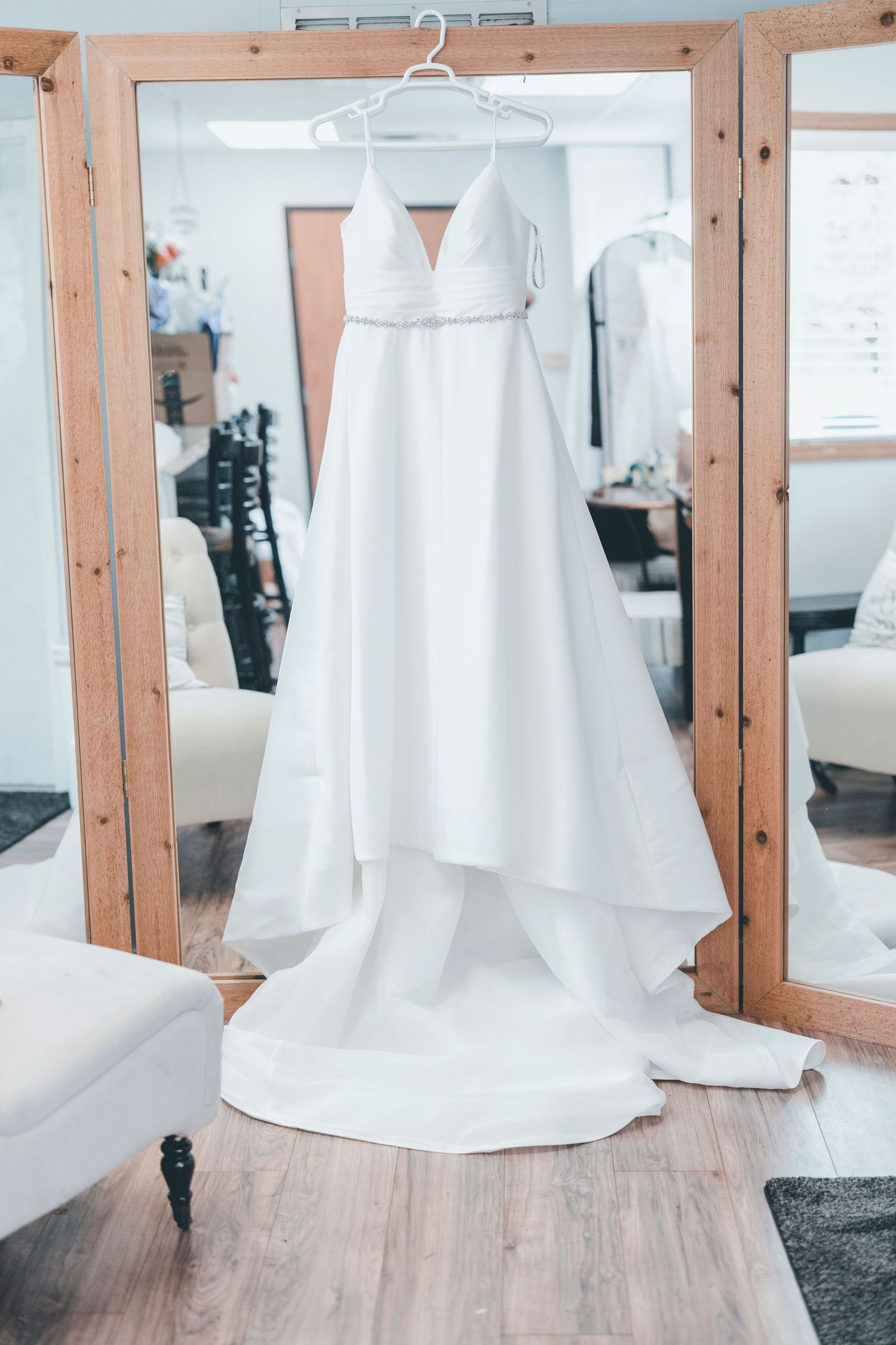A white wedding dress hangs on a wooden hanger against a light-colored wall with exposed wooden beams.
