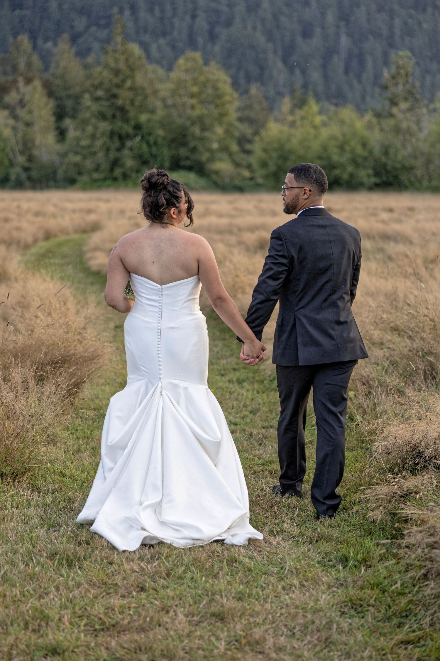 Newlyweds walking through a grassy mountain field with evergreen trees in the background.