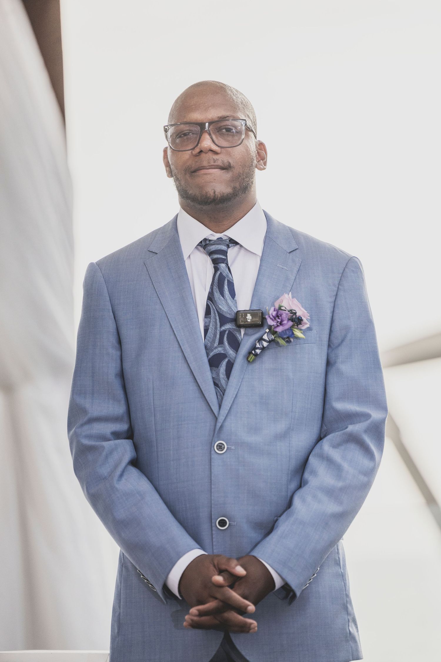 Groom in light blue suit with glasses and boutonniere smiles while standing against white background at wedding.