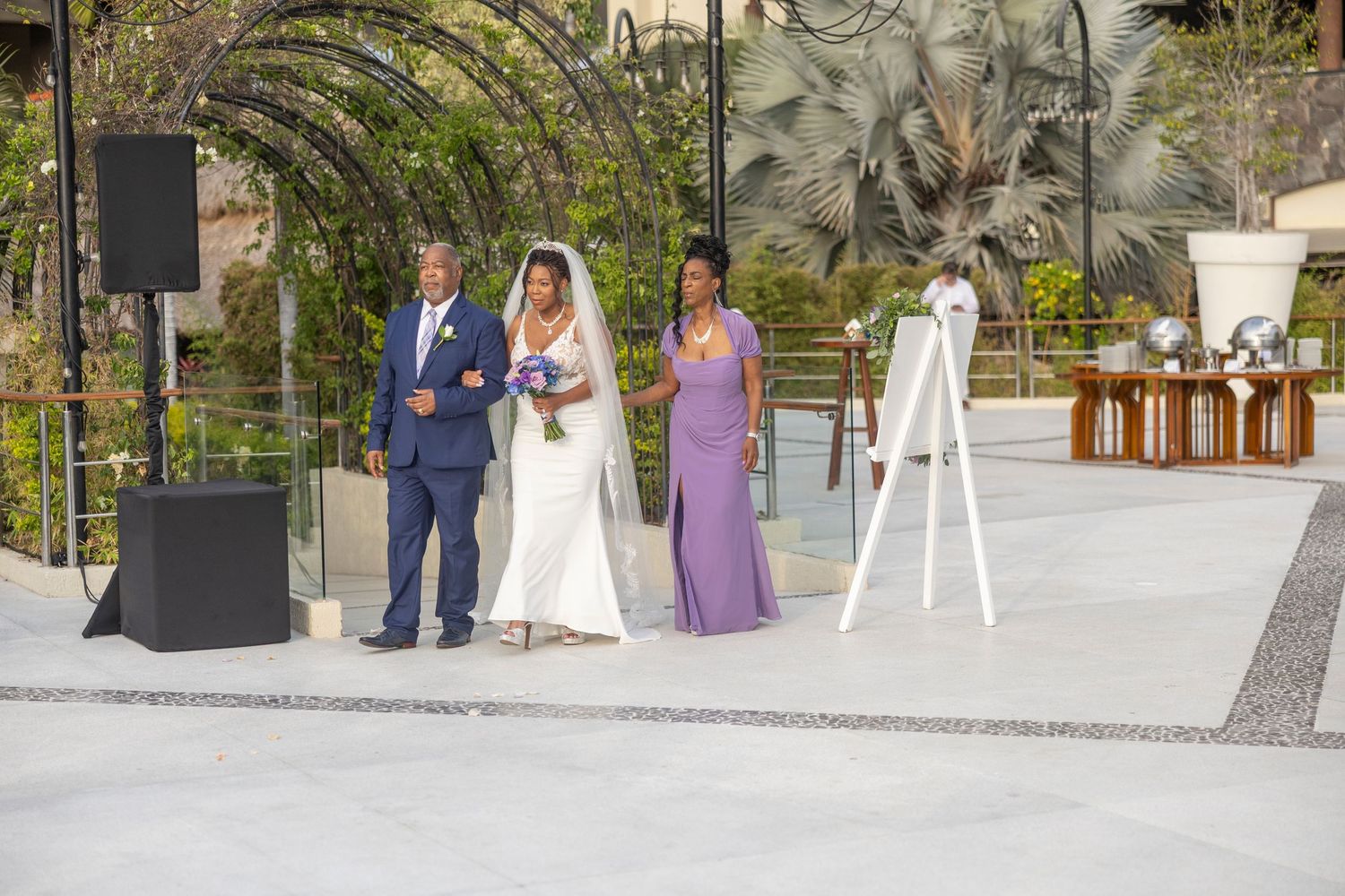 Wedding party stands in a garden courtyard during an outdoor ceremony with palm trees and decorative elements.
