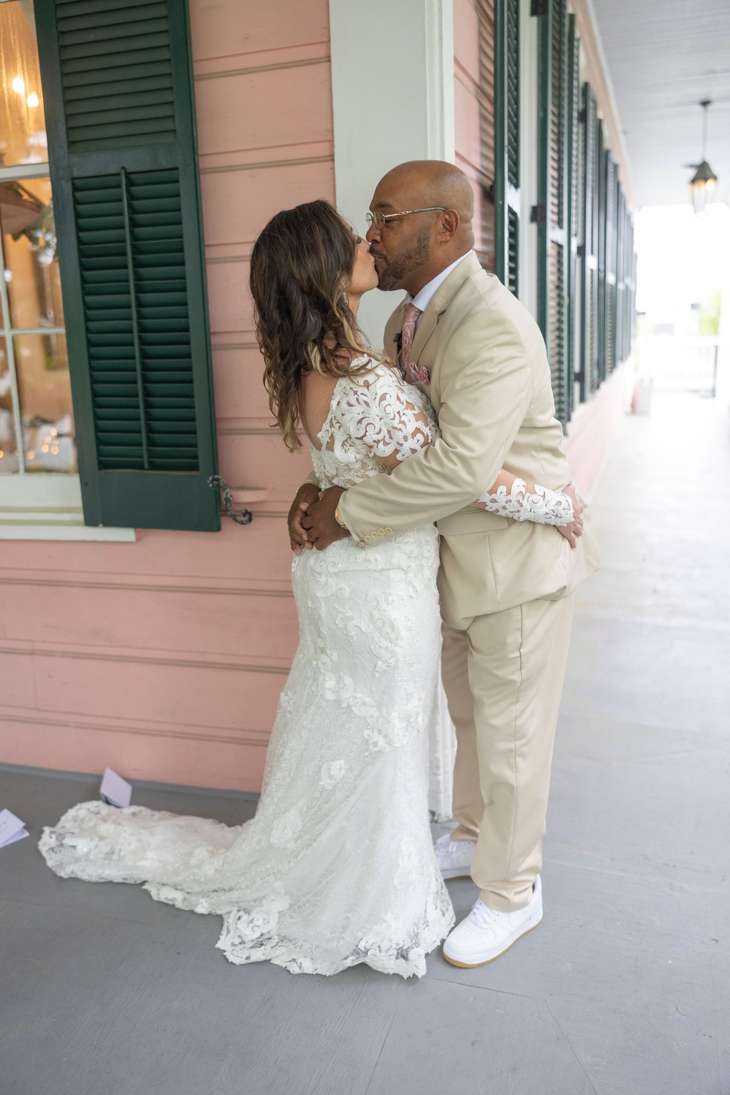 A couple in wedding attire shares a tender moment on a porch with green shutters and pink walls.