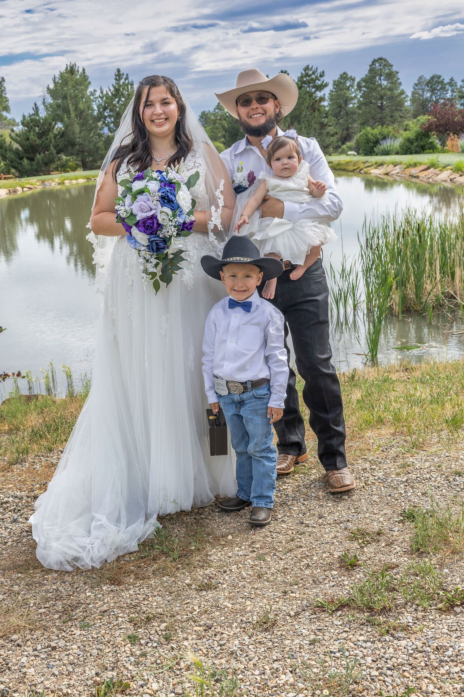 A wedding party poses together by a serene lake with white dresses and cowboy hats on a sunny summer day.