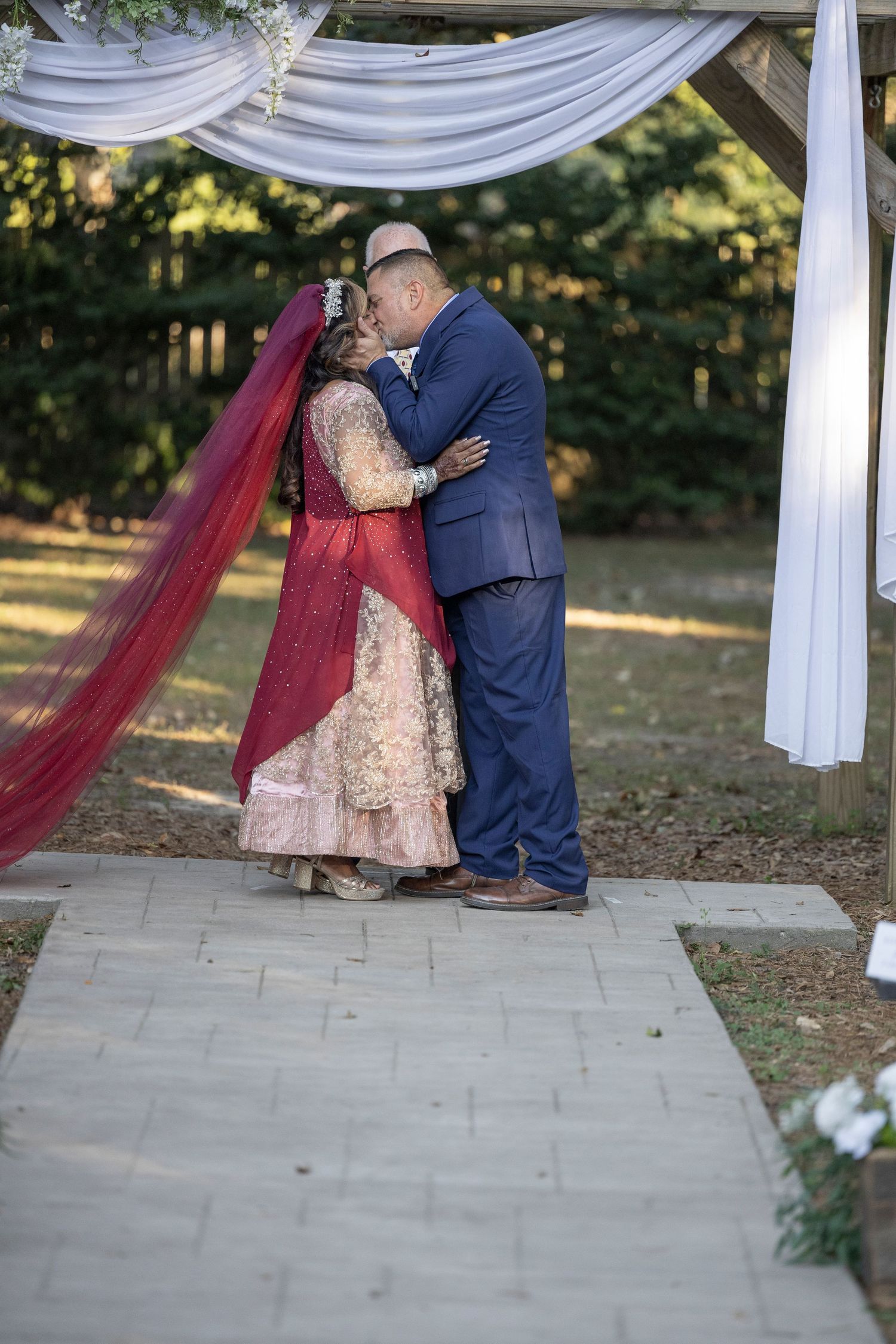 A couple shares an intimate moment under a white draped wedding arch while wearing traditional South Asian wedding attire.