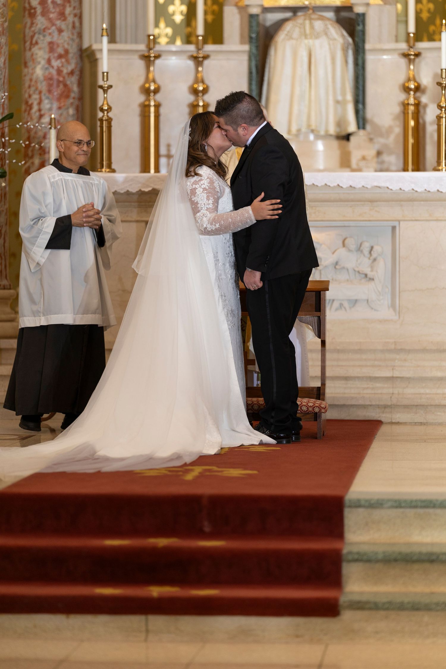 Wedding ceremony taking place inside an ornate church with white and gold decor and red accents on the walls.
