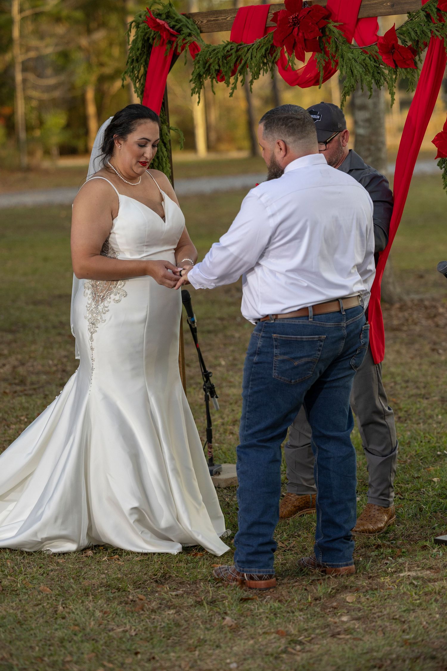 A couple in wedding attire exchanges rings during an outdoor autumn ceremony beneath red ribbons and evergreen branches.