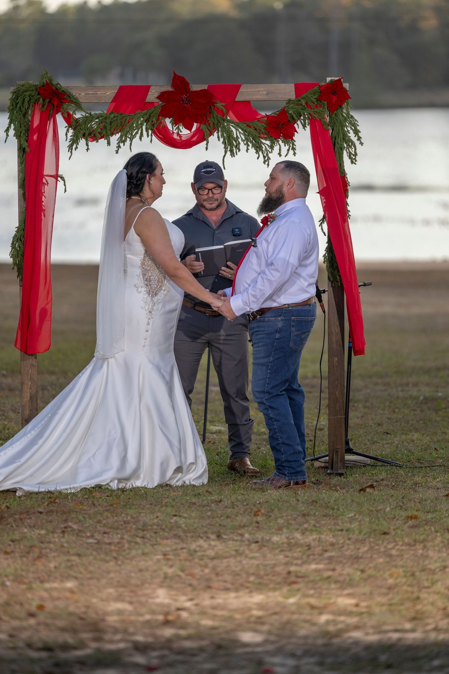 Wedding ceremony takes place outdoors under a rustic wooden arch decorated with red ribbons and greenery by a lake.